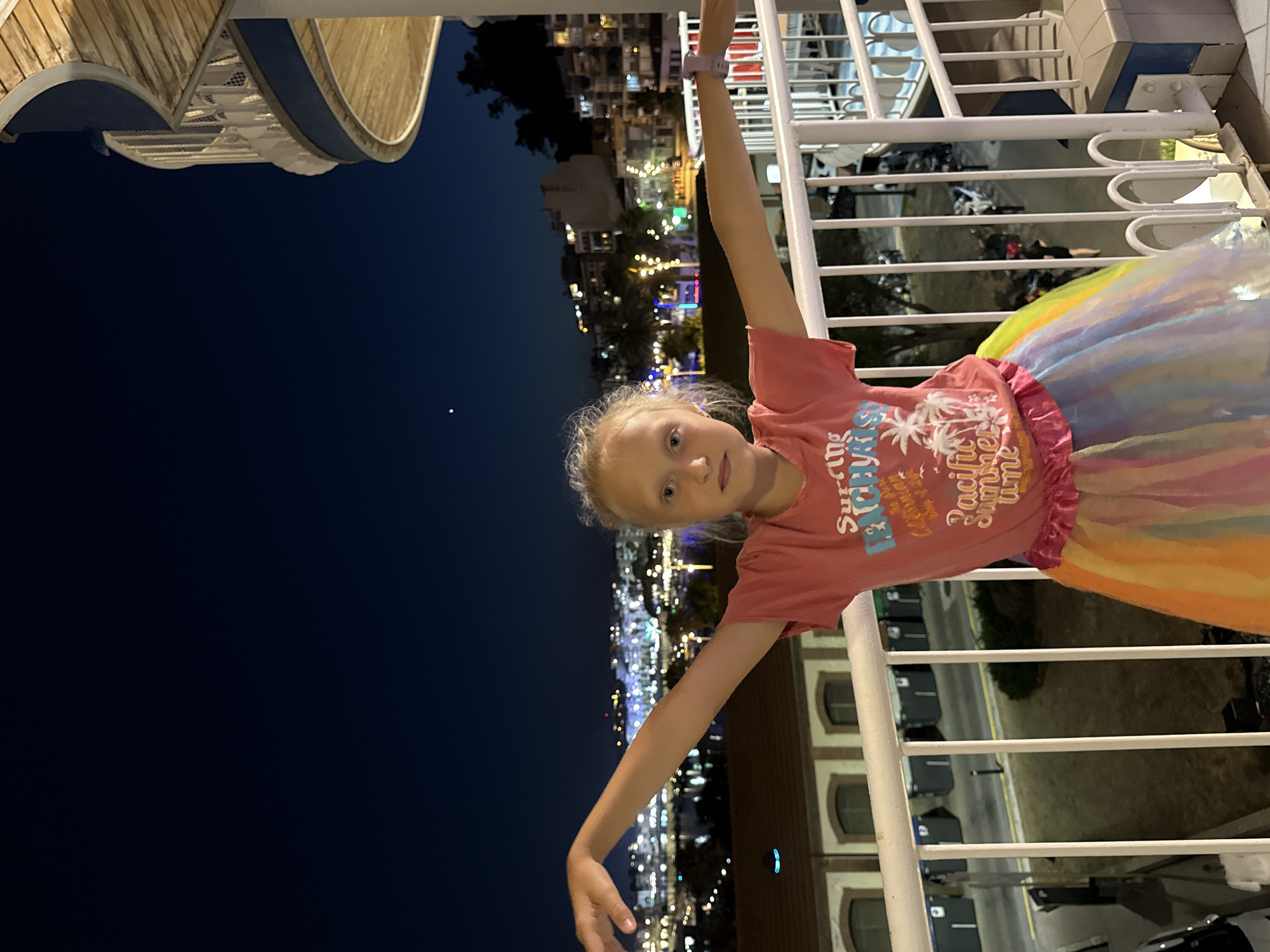 A young girl with blonde hair stands on a balcony at night, extending her arms outwards. She is wearing a pink t-shirt with graphics and a multi-colored, iridescent skirt. Behind her, the city of Blanes, Spain, is illuminated with many lights, extending up a hillside. The sky is dark blue, with a single bright star visible. A spiral staircase structure is partially visible on the right side of the image. The balcony has a white railing and a concrete floor. The building in the background appears to be an apartment complex or hotel.