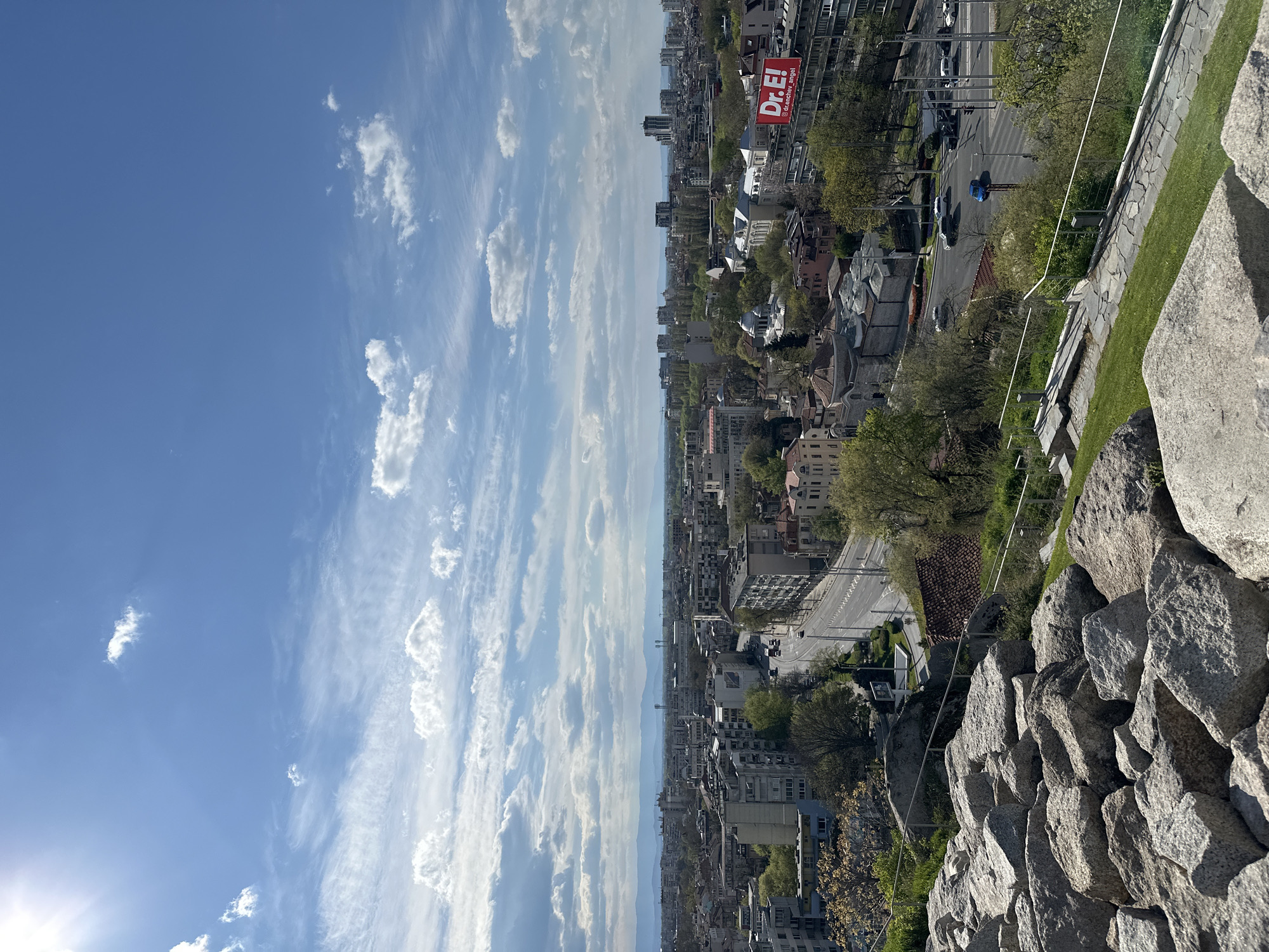 The image shows a cityscape view from a high vantage point in Plovdiv, Bulgaria. The sky is blue with scattered cumulus clouds. Below, a densely populated urban area unfolds with buildings of varying heights and architectural styles, interspersed with green spaces and trees. A street with light traffic is visible, and a red billboard with the word "Dr. E!" is prominently displayed on one of the buildings. In the foreground, a stone wall with large rocks forms the edge of the viewing area, indicating the elevated position. A person in blue is visible walking on the street. The sun is shining brightly from the lower left corner.