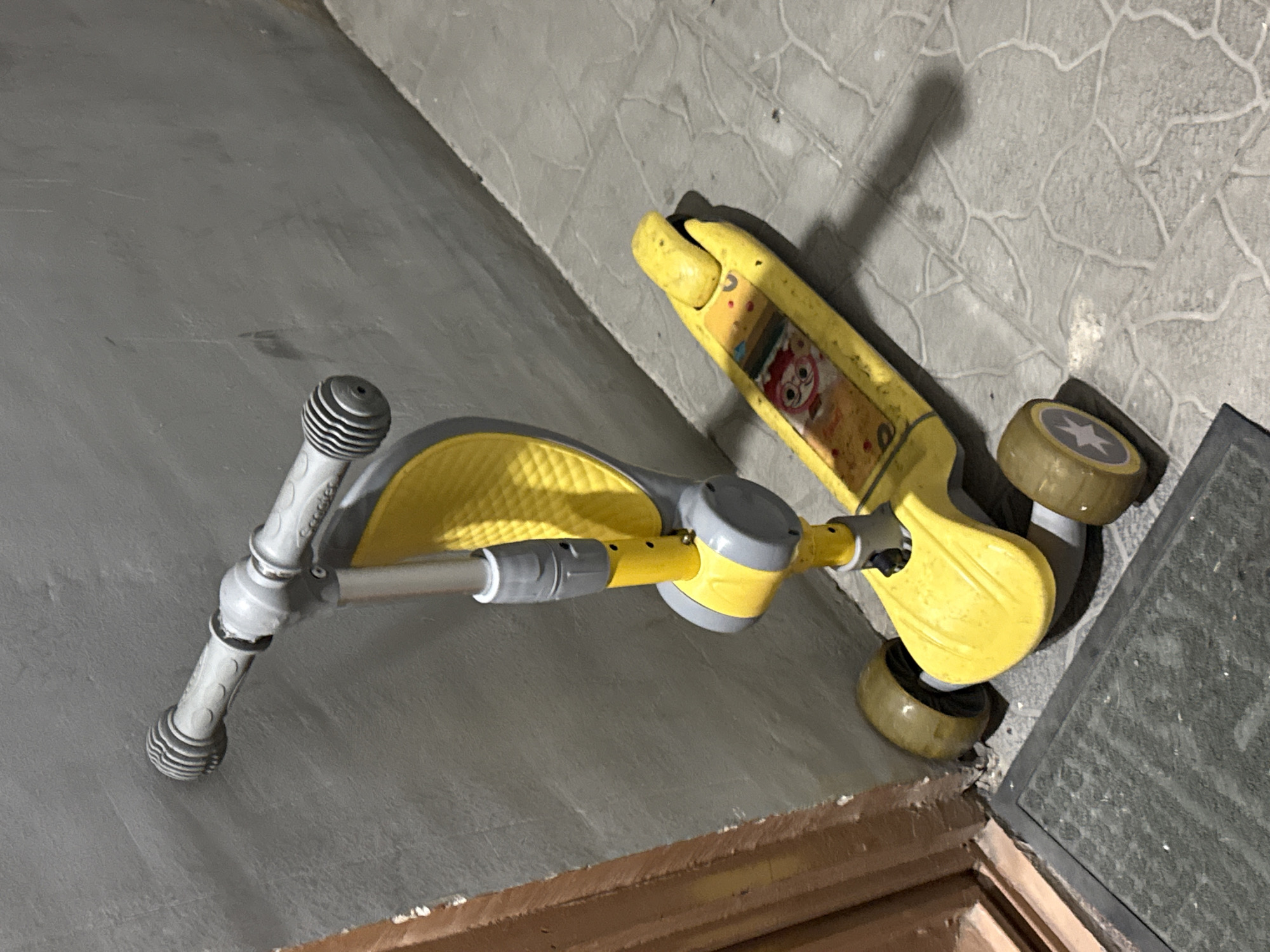 An outdoor shot depicts a yellow and gray three-wheeled scooter leaning against a textured gray wall. The scooter is positioned to the right of a dark brown doorframe and a dark gray doormat. The scooter has yellow handles, a yellow deck with a cartoon character sticker, and yellow and gray wheels. The scooter is stationary and no people are present. The ground is paved with gray tiles. The lighting suggests it is nighttime. The scene is located in Tbilisi, Georgia.