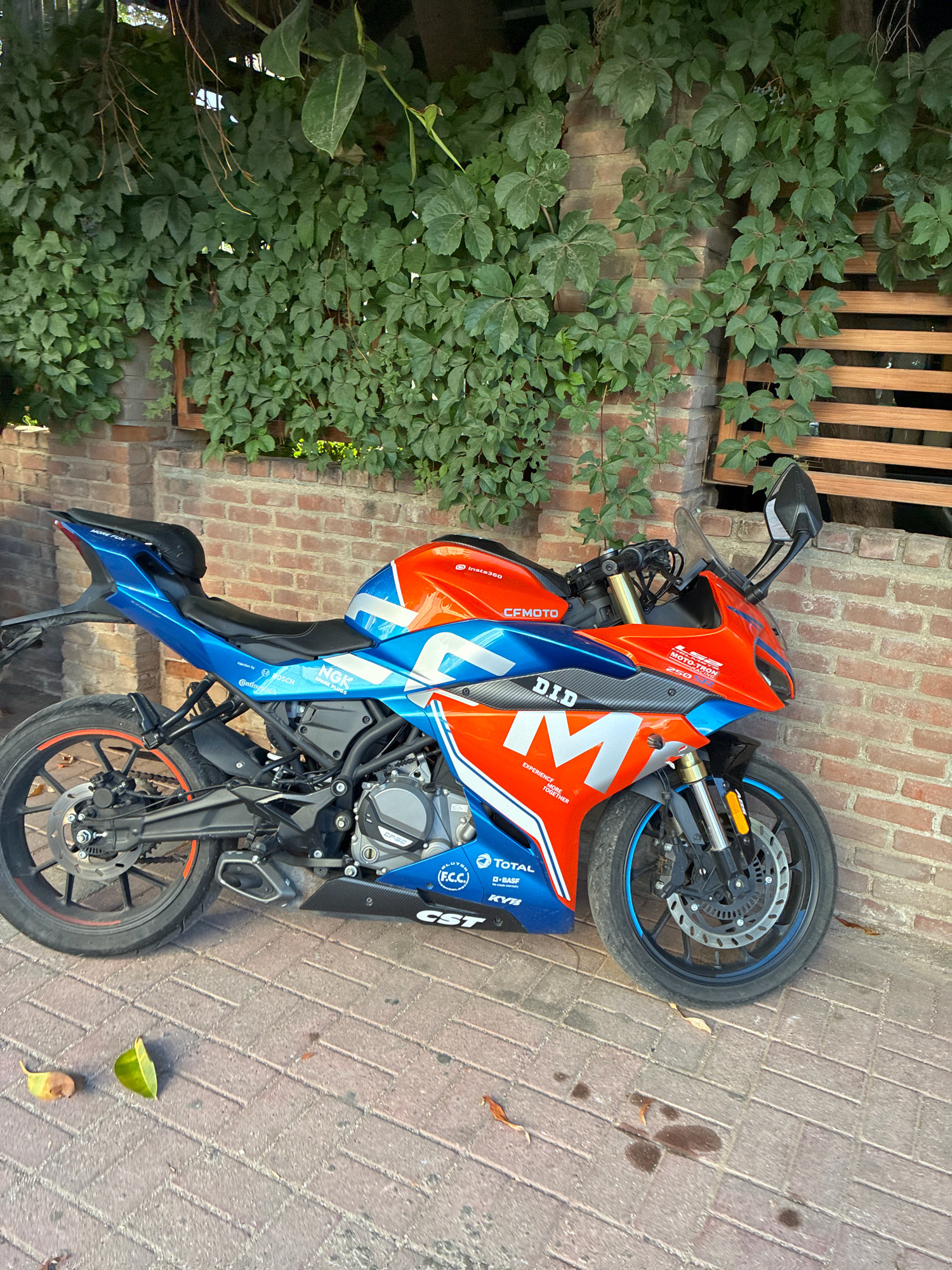 An orange, blue, and silver sport motorcycle is parked on a brick-paved walkway next to a brick wall covered in ivy. The motorcycle has various logos and text printed on its fairings, including "CFMOTO," "DID," and "TOTAL." A small black bag is attached to the rear seat. Two fallen leaves are on the walkway in front of the motorcycle. In the background, a wooden slatted structure is visible. The scene appears to be outdoors, likely in an urban or town setting.