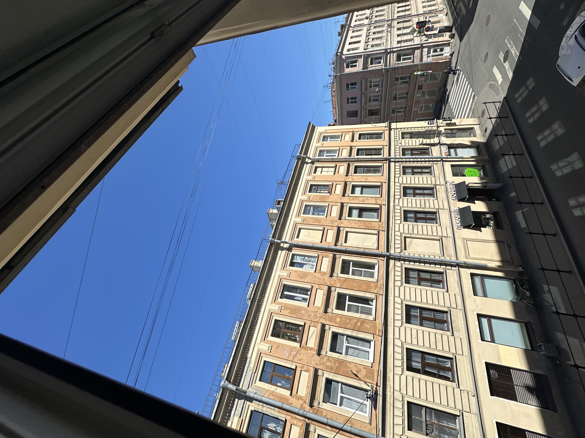The image presents an elevated view overlooking an urban street intersection on a clear, sunny day. A vast, cloudless blue sky occupies the upper frame, intersected by multiple parallel utility wires.
In the mid-ground, a multi-story building with a weathered, light ochre and pale yellow facade dominates the left side. It features numerous rectangular windows, some with dark brown frames and others with white modern frames; some windows are open, while others display curtains or blinds. Architectural details include decorative cornices and window surrounds. Two grey drainpipes are affixed to its front. The ground floor houses commercial spaces, one identified by Cyrillic text "ПЕКАРНЯ КОФЕЙНЯ" (Bakery Coffee Shop) above a dark awning, with a bright green apple-shaped sticker on an adjacent window. A bicycle is parked against the building near a storefront.
To the right and further in the background, another multi-story building with a light pink facade is visible, also featuring numerous windows and architectural details typical of European urban centers.
The asphalt street below forms a clear intersection marked with white zebra crosswalks. Traffic lights are present at the corner. Bright sunlight casts sharp, elongated shadows of architectural elements, a metal railing, and overhead wires onto the street, indicating intense daytime illumination. A black metal railing runs along the foreground sidewalk.
One pedestrian is visible crossing the street at the intersection. A white car is partially visible in the bottom right corner of the frame. The location is an unknown city, in an unknown country.