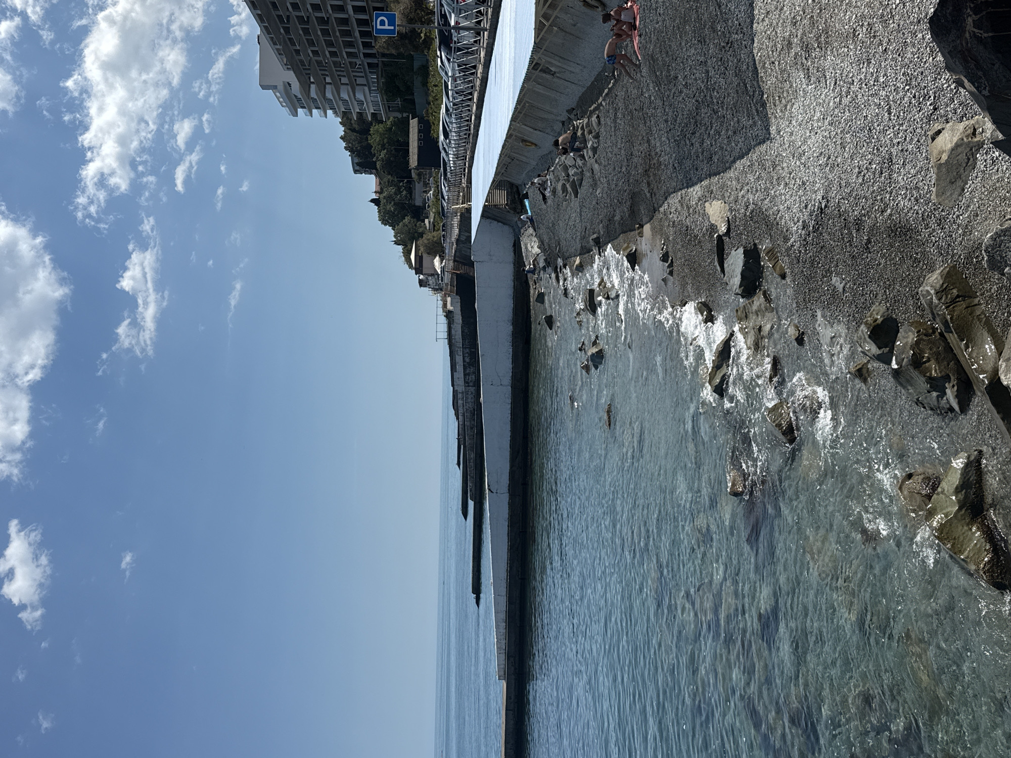 The image displays a bright, sunny coastal landscape viewed from an elevated vantage point. A clear blue sky with scattered white cumulus clouds fills the left side of the frame. The sea, a translucent blue-green, occupies the middle ground, with small waves gently breaking against the shore.
Along the right side, a dark, rocky and pebbly beach extends into the distance. A long, white concrete sea wall or promenade runs parallel to the coastline, separating the beach from an area further inland. Several large, dark rocks are scattered along the water's edge and within the shallow sea. A few people are visible on the rocky shore; two individuals are lying or sitting on towels near the upper part of the beach, while another person appears to be crouching closer to the water.
Above the promenade, a road is visible with several cars parked along its side. Beyond the road, multi-story buildings with numerous windows and balconies ascend the hillside, interspersed with green trees and foliage. A blue rectangular sign with a white capital "P" (indicating parking) is prominently displayed near one of the buildings. The bright sunlight and calm waters suggest a pleasant daytime setting, likely during good weather.
Location context: Unknown, Unknown