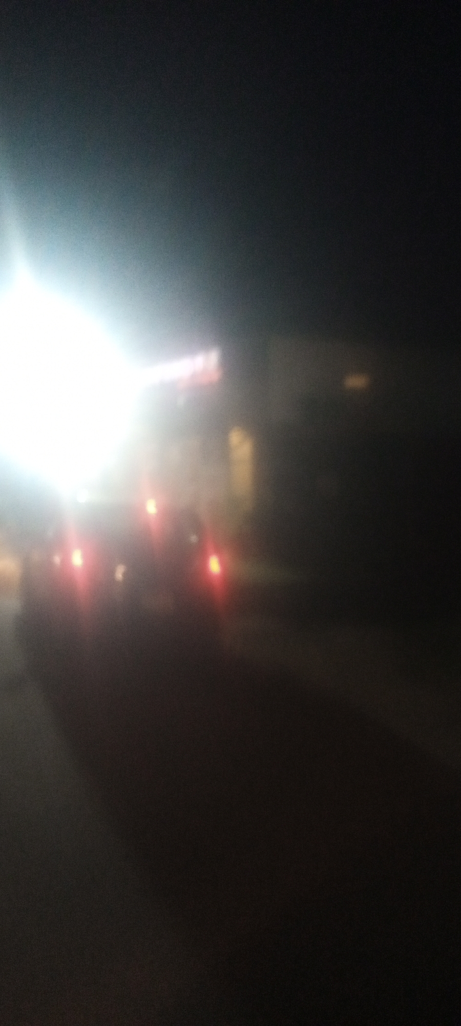 The image shows a dark, blurry scene at night, likely in Narayi, Nigeria. A car is visible with its headlights and taillights on, suggesting it is either approaching or stationary with its rear facing the camera. In the background, there is a building or structure with some illuminated signage or windows. The overall environment appears dark and visibility is limited. There are no other discernible people or objects visible.