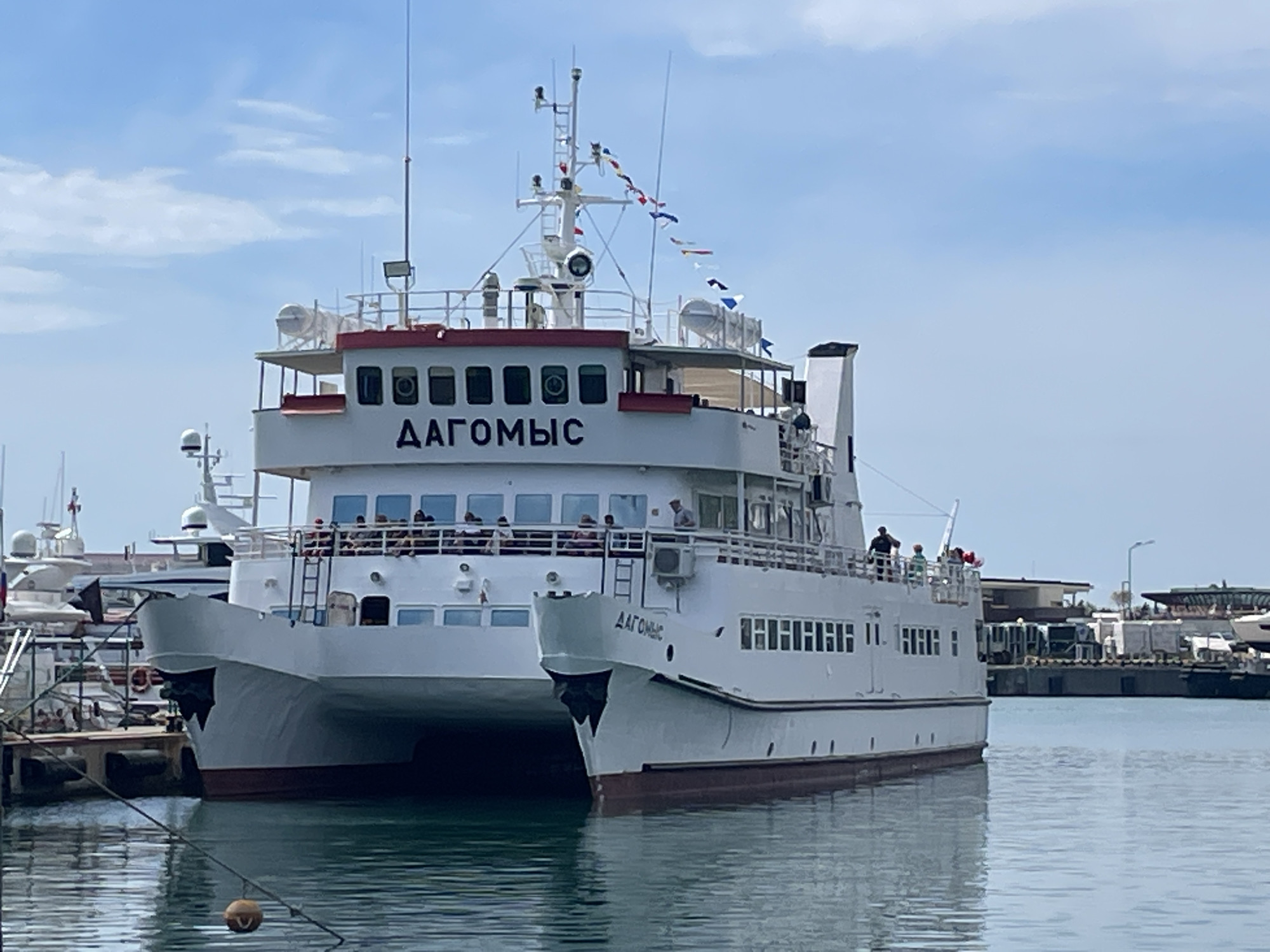 The image shows a white catamaran ferry docked in a harbor. The ferry has a red trim and is named "ДАГОМЫС" in Cyrillic letters on its side. There are people visible on the upper deck of the ferry. In the background, other boats and port facilities are visible, suggesting a busy harbor environment. The sky is partly cloudy and the water is calm. The location is the port of Dagomys in Sochi, Russia, a coastal city on the Black Sea known for its resorts and beaches.