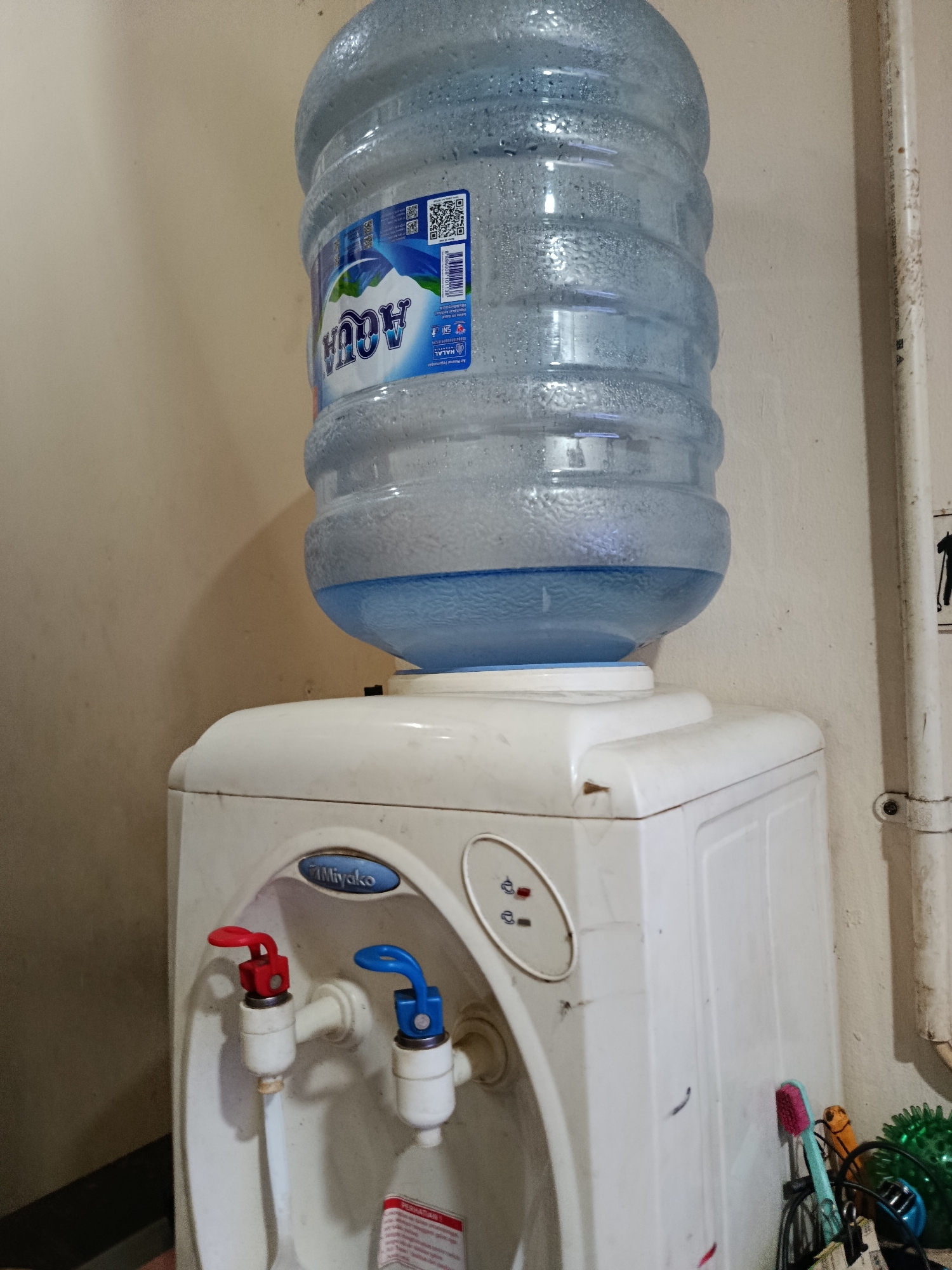 The image shows a close-up of a water dispenser with a large bottle of Aqua brand water on top. The dispenser is white and has two spigots, one red and one blue, indicating hot and cold water respectively. A brand name "Miyako" is visible on the dispenser. The background is a plain, light-colored wall with some pipes running vertically. There are also some cleaning supplies and a green spiky object to the right of the dispenser. The overall scene suggests a domestic or office setting. The location is not specified in the image, so it cannot be determined to be in Central Jakarta, Indonesia.