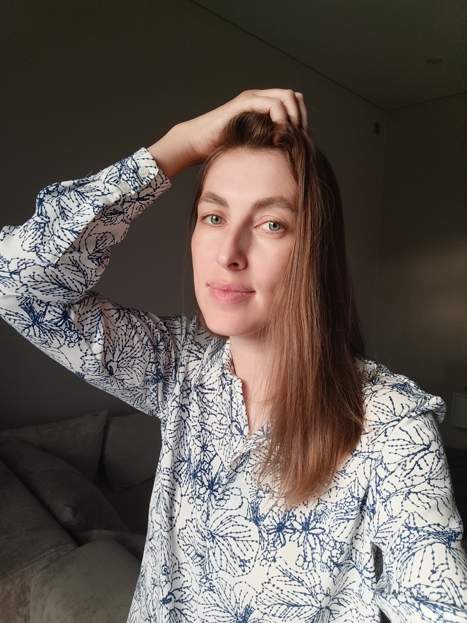 A young woman is shown from the chest up, looking at the camera. She has long, light brown hair and is wearing a white blouse with a blue floral pattern. Her right arm is raised, with her hand in her hair. The background is dark, with the exception of a couch on the left side of the frame. The lighting suggests the sun is casting shadows. There is no information to determine the location in the city of Chaikovsky, Russia.