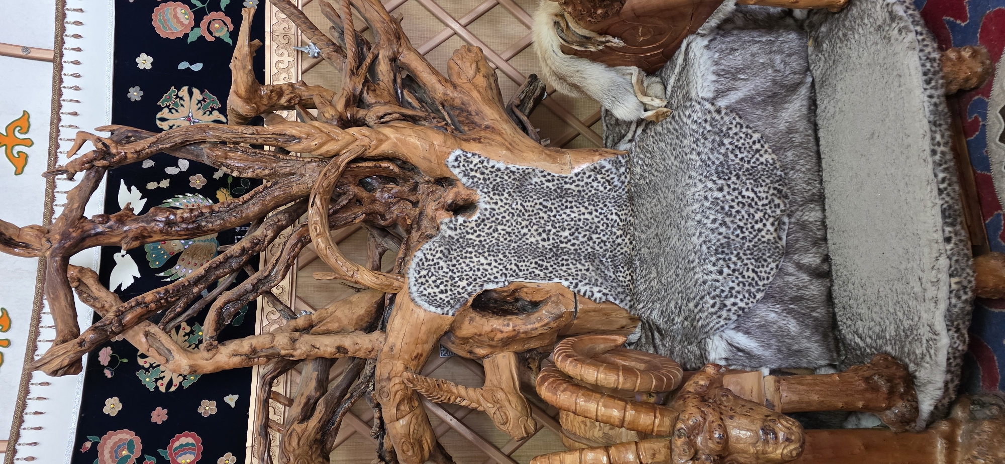 This is a full-frame, eye-level, medium shot of a decorative throne-like chair. The chair appears to be made of intricately carved wood, resembling tree branches and roots, with animal hides serving as upholstery. The main structure of the chair is a large, gnarled wooden piece that forms the back and arms, with the wood displaying a natural, unfinished texture.
The seat and backrest of the chair are covered with animal furs. A spotted hide, possibly leopard print, is draped over the back and part of the seat, while a more grey and white mottled fur covers the main seating area. The left armrest is carved to resemble the head and horns of a ram. A fox pelt is draped over the right armrest, with its head and tail visible. The base of the chair rests on wooden legs.
Behind the chair, a patterned fabric hangs on the wall, featuring floral motifs and depictions of birds. The environment appears to be indoors, possibly within a dwelling or a cultural exhibition space, given the decorative elements and the construction of the chair. The lighting is even and natural, suggesting daytime. No people are present in the image. The overall impression is one of a rustic, perhaps traditional, seating arrangement with a focus on natural materials and animal motifs. The context of Bulan-Segettu, Kyrgyzstan, suggests a potential connection to nomadic or traditional Kyrgyz culture.