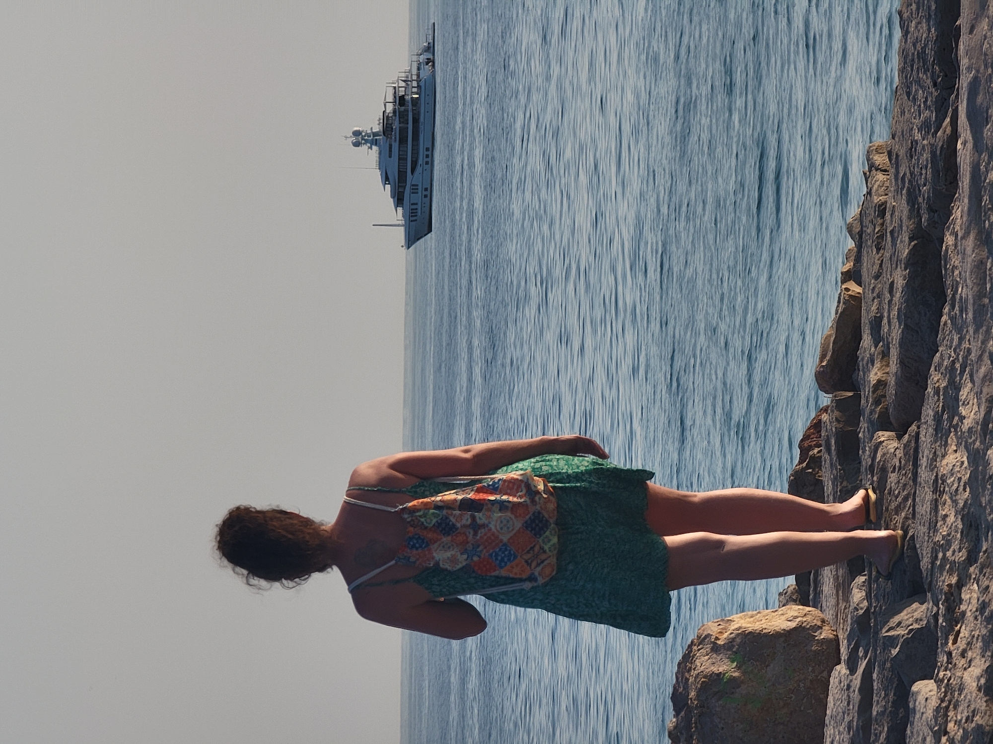 A woman stands on a rocky shoreline, facing the sea. She is wearing a green, knee-length sundress and a colorful, patchwork backpack. Her dark, curly hair is pulled up into a bun. In the distance, a large white yacht is visible on the water. The sky is a pale, hazy gray. The woman is standing on Quarteira's breakwater, a prominent feature of the city's coastline.