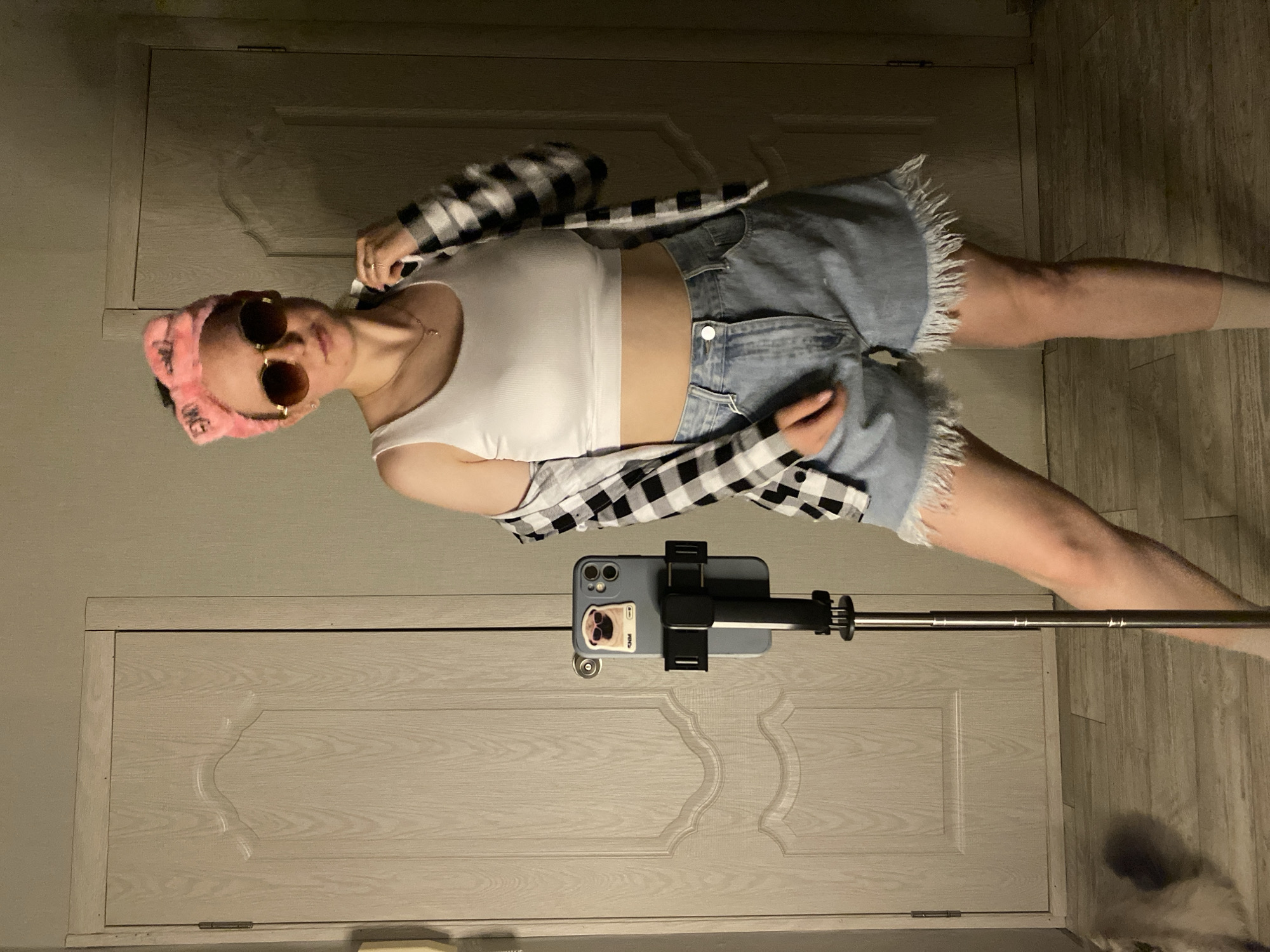 An individual is depicted standing indoors in an unknown location in an unknown city. The person, appearing female with light skin and dark hair partially obscured by a pink furry headband, wears a white ribbed crop top, light blue distressed denim shorts with frayed hems, and an open black and white plaid shirt. She also wears round mirrored sunglasses, a gold necklace with a small pendant, and light-colored socks. Her right hand is near her left collarbone, touching the plaid shirt. To her left, an iPhone, encased in blue and featuring a pug sticker, is mounted on a black and silver extendable tripod. The background consists of two light-colored, paneled wooden doors and a light-colored wooden plank floor. The walls are plain and light-toned. Lighting is diffused, creating subtle shadows. A blurry, light-colored object is visible on the floor in the lower-left corner.