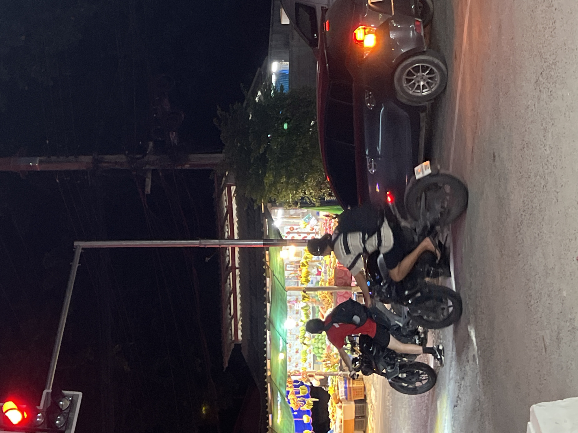 This is a night-time, street-level photograph taken in the Ko Pha-ngan District of Thailand. The scene depicts a road intersection with a traffic light showing red. Two individuals on motorcycles are stopped at the intersection. The motorcycle on the left is ridden by a person wearing a red t-shirt and black shorts, carrying a black backpack. The motorcycle on the right is ridden by a person wearing a striped t-shirt and dark shorts, also carrying a black backpack. Behind the motorcycles, a dark-colored car is stopped, with its brake lights illuminated. In the background, there is a market stall with illuminated displays of fruits. The overall environment suggests a commercial area on an island.