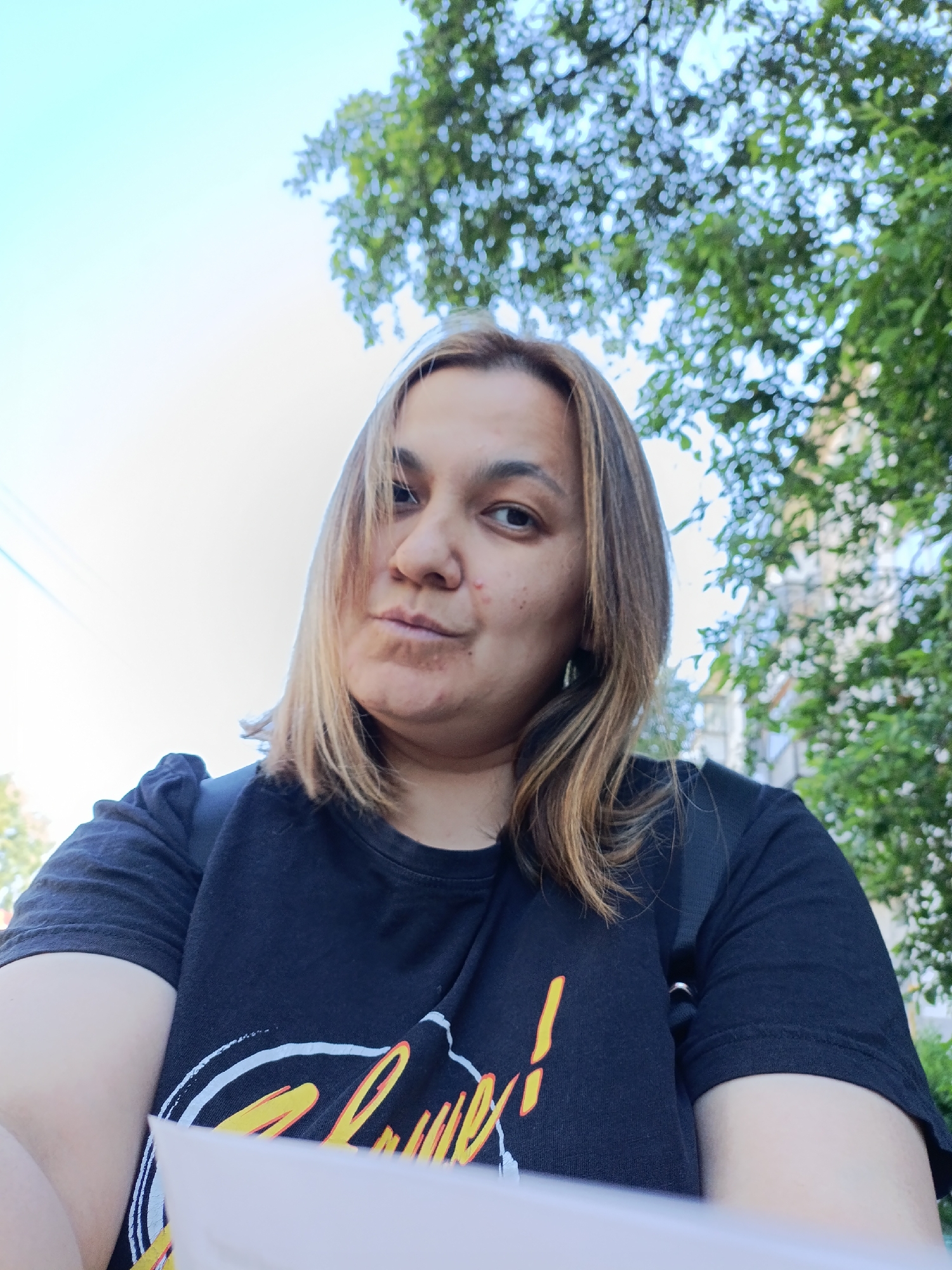 A selfie taken outdoors features a woman with shoulder-length brown hair with blonde highlights, facing the camera with a neutral expression. She is wearing a black t-shirt with a graphic design. The background consists of lush green foliage and a bright sky, suggesting a daytime setting. The image appears to be taken in an urban environment, possibly a park or residential area, with the presence of buildings and power lines subtly visible. The woman is holding a white piece of paper in the foreground.