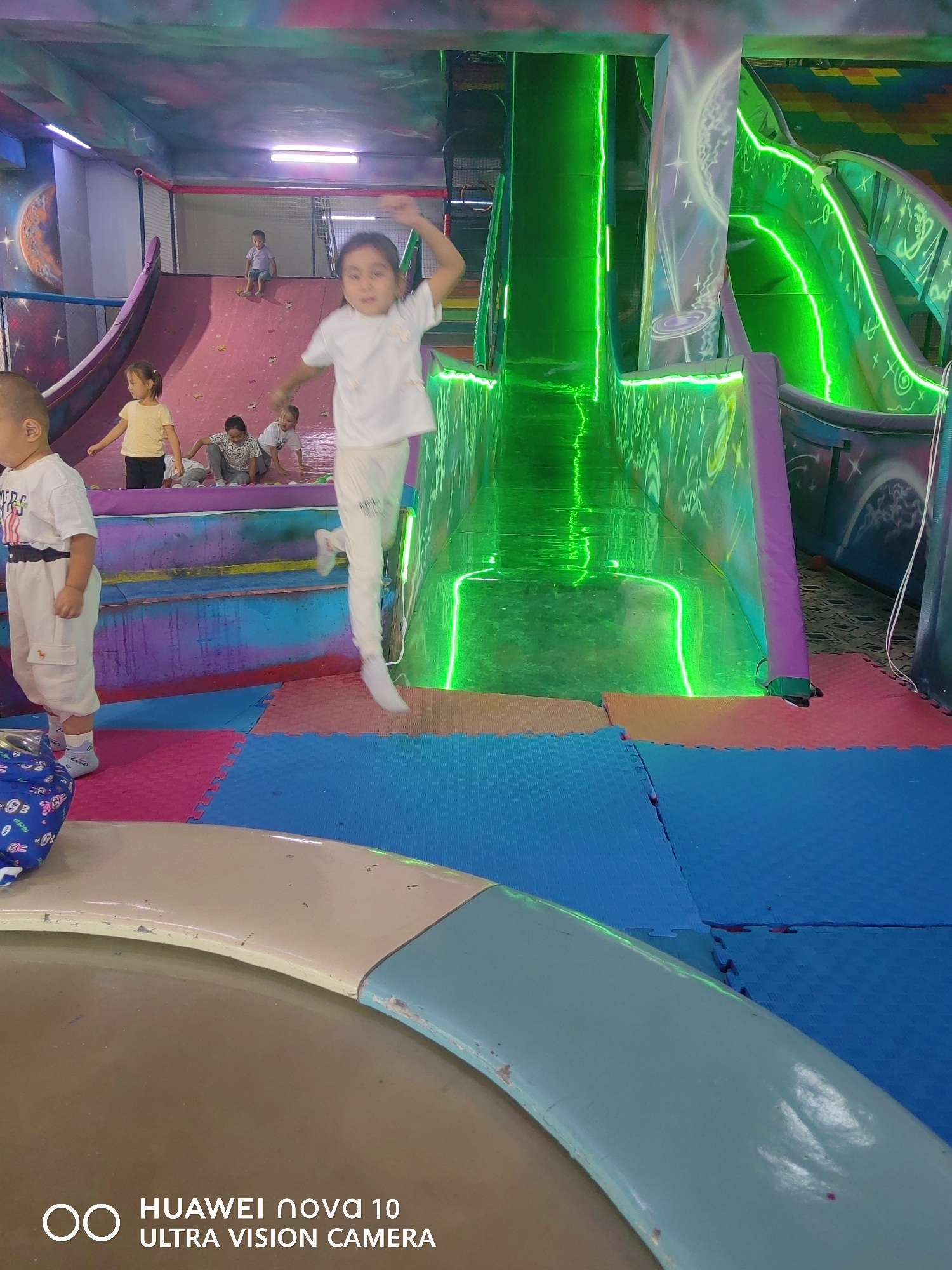The image depicts an indoor children's play area, likely a soft play center or amusement park, with a focus on a vibrant, neon-lit slide system. In the foreground, a child in white pants and a white t-shirt is captured mid-jump, with their arms outstretched. To the left, another child, also in white, stands looking towards the camera. Behind them, several other children are visible, some sitting on a pink climbing wall and others on the padded floor. The environment features brightly colored, interlocking foam mats on the floor in shades of blue, pink, and orange. The play structure includes a large, tubular slide that glows intensely with green neon lighting, creating a futuristic and energetic atmosphere. The walls and ceiling are painted with whimsical designs, including planets and stars, further enhancing the playful theme. The overall impression is one of active engagement and fun within a dedicated children's entertainment space. The image metadata indicates that the location is Özgön, Kyrgyzstan.