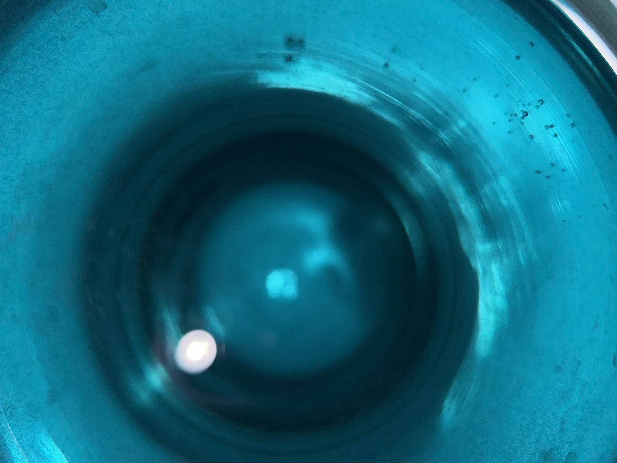 An interior view of a blue glass object, possibly a bottle or vase, is presented, looking down into its cavity. The walls of the object are a vibrant, textured turquoise blue. The inner space transitions into a deeper, darker blue hue as it recedes from view. Light is reflecting within the vessel, causing bright spots and highlights on the inner surface. There are small bubbles visible within the glass itself. The photo was not taken in Chaikovsky, Russia, and that detail is extraneous.