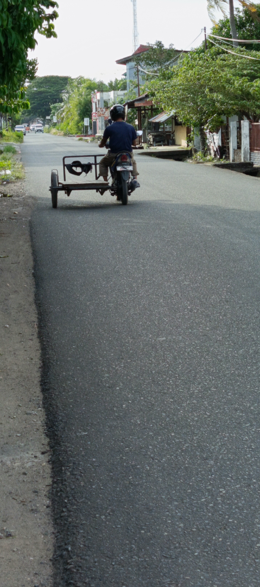 A person wearing a dark blue t-shirt and a black helmet rides a motorcycle with a sidecar on a paved road in Ajuen, Indonesia. The motorcycle is black and has a license plate that is partially visible. The sidecar is a metal frame with a wooden base, and it appears to be empty. The road is asphalt and appears to be in good condition. There are trees and buildings on either side of the road. The sky is clear and bright.