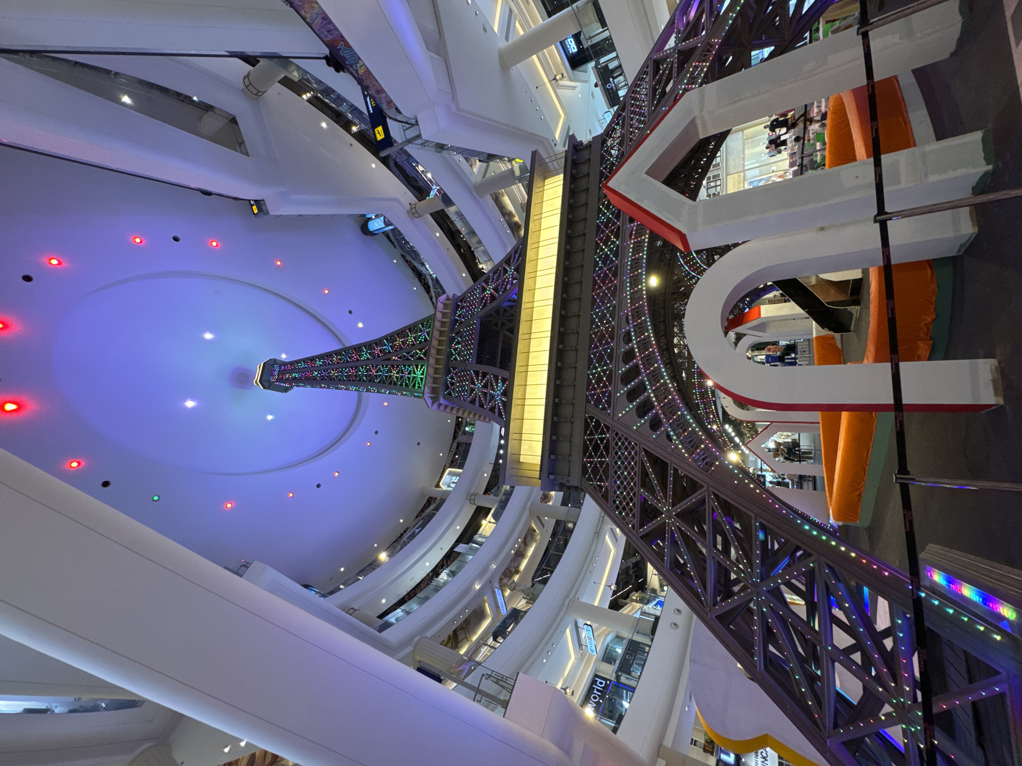 An indoor shot, taken from a low angle looking up at a miniature Eiffel Tower replica adorned with colorful lights, stands prominently in the center of a multi-level shopping mall atrium. The replica is positioned above a play area with orange inflatable structures and white archways. Surrounding the central display are multiple floors of the mall, featuring balconies with shops and escalators. The mall's ceiling is a large, domed structure illuminated with blue and red lights, creating a vibrant atmosphere. The scene is located within Terminal 21 Pattaya, a themed shopping mall in Pattaya City, Thailand, known for its "world city" concept.