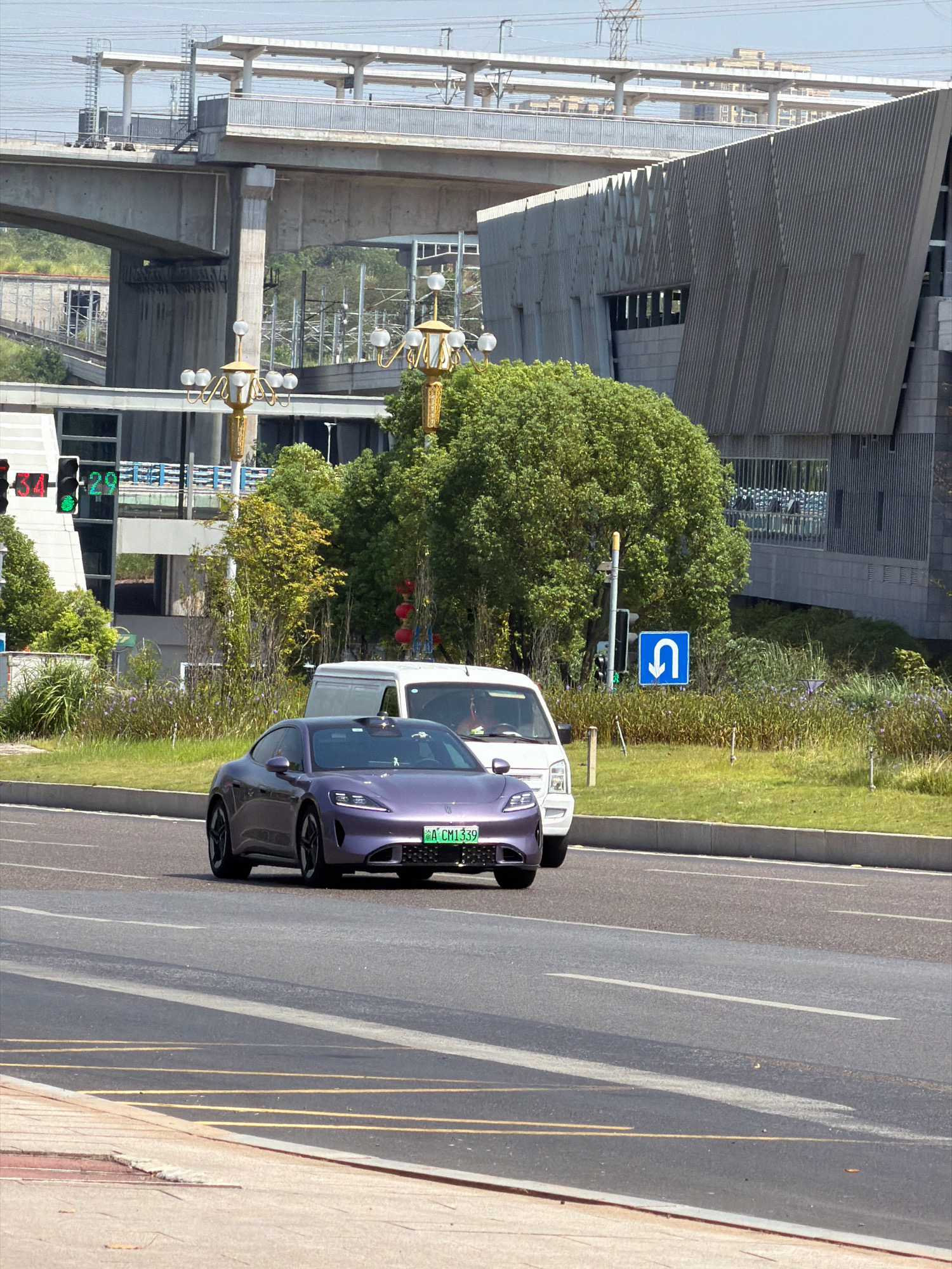 In Jiangjin, China, a purple electric car, identified by the license plate "A CM1339", drives on a paved road. Behind the car is a white van. The scene features an elevated concrete structure, likely part of an overpass or transit system, with overhead wires. A modern building with a textured grey facade is visible, alongside lush green trees and shrubs. A traffic light shows a green signal, and a blue sign with a white U-turn arrow is posted. The area appears to be a well-maintained urban environment.