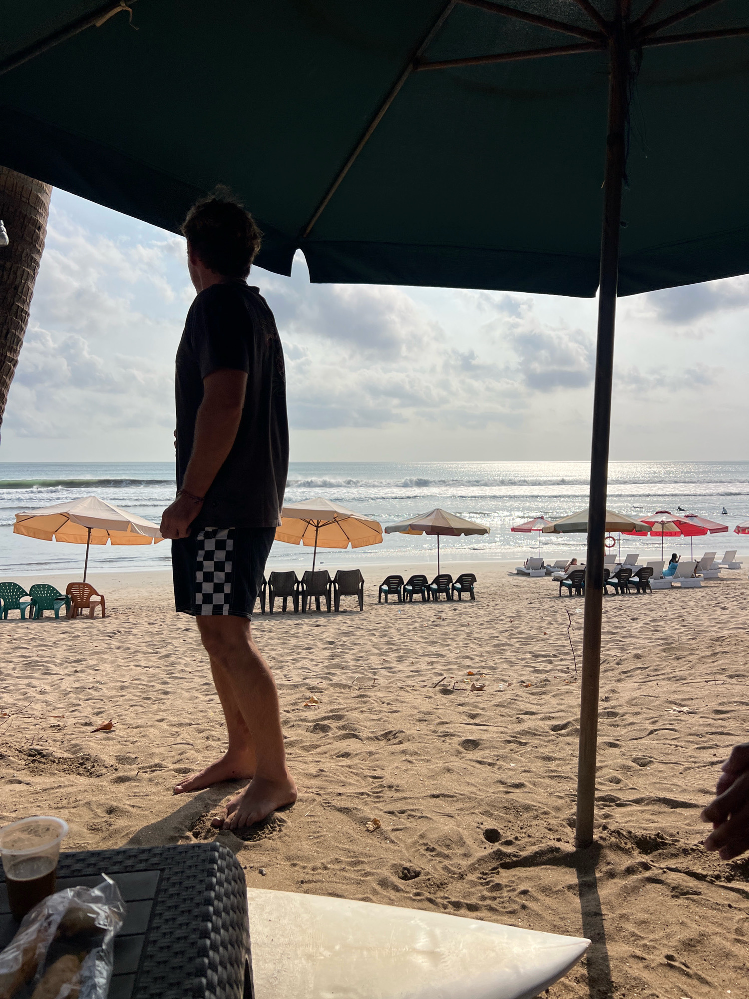 A man stands on a sandy beach in Kuta, Indonesia, facing the ocean. He is wearing a dark t-shirt and checkered shorts. In the foreground, a dark woven table has a plastic cup with a dark liquid and a plastic bag with food items on it. A portion of a white surfboard is visible at the bottom right of the frame. Several beach umbrellas and chairs are set up on the sand. The ocean has moderate waves, and the sky is partly cloudy with the sun glinting on the water. The overall scene depicts a relaxed beach atmosphere.