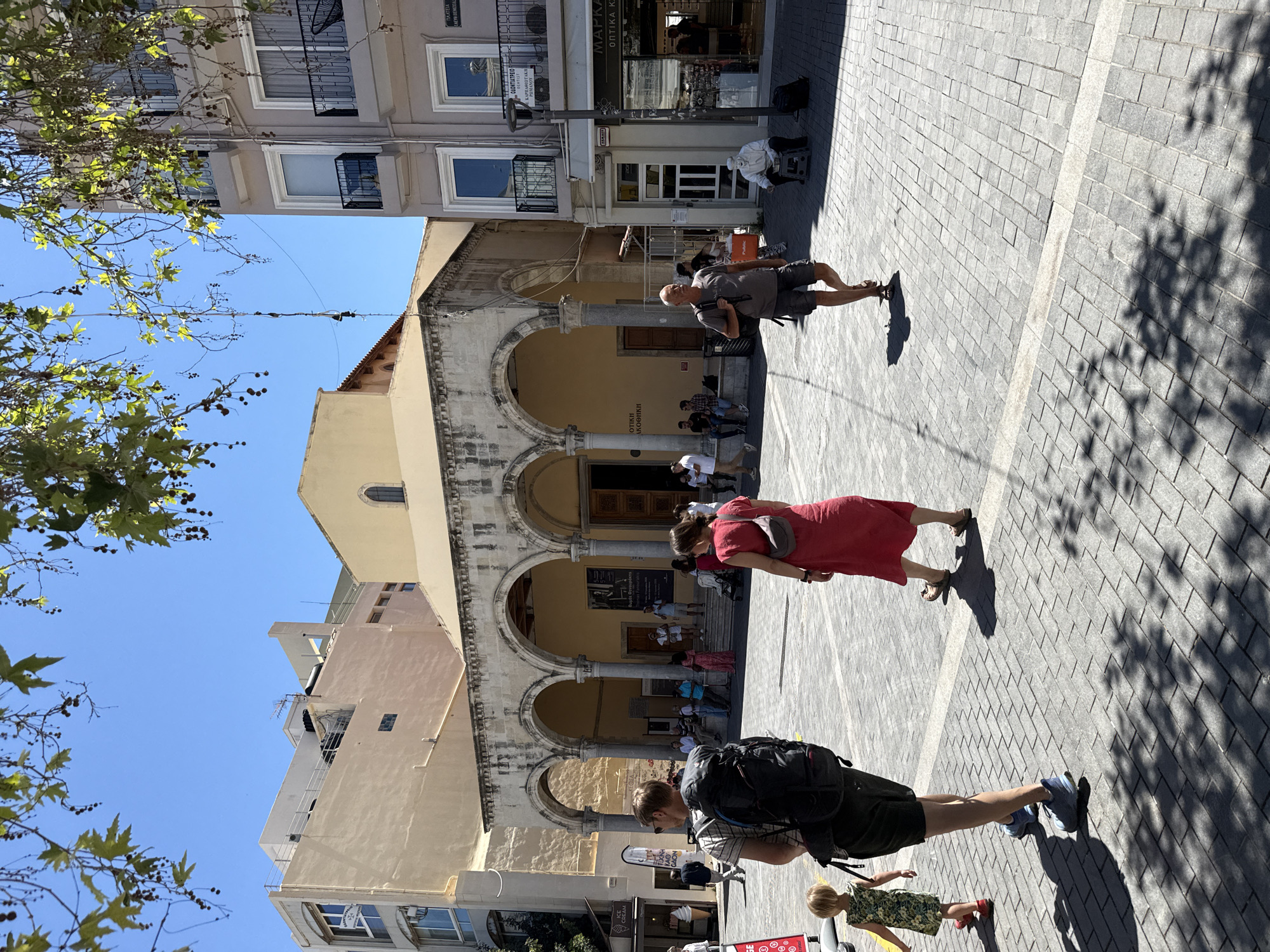 A sunny daytime scene unfolds in a public square paved with light gray stone tiles, likely in Heraklion, Greece. A large building with arched walkways and a pale yellow facade dominates the background, suggesting a historical or religious structure. Adjacent to it is a modern, multi-story building with balconies and windows.
In the foreground, several individuals are navigating the square. A man with a large black backpack is walking away from the viewer, accompanied by a young child in a floral dress. To their right, a woman in a vibrant red dress walks past. Further to the right, a man in a grey t-shirt and shorts walks towards the viewer, carrying a shopping bag. Near the building on the right, an elderly person dressed in white is seated on a stool, appearing to be a street performer.
The square is shaded in parts by the foliage of trees overhead, casting dappled patterns on the ground. Signs are visible on some of the storefronts, including one for "MARKASI OPTIKA KAO", indicating shops are present. The overall atmosphere suggests a lively urban environment during daylight hours.
