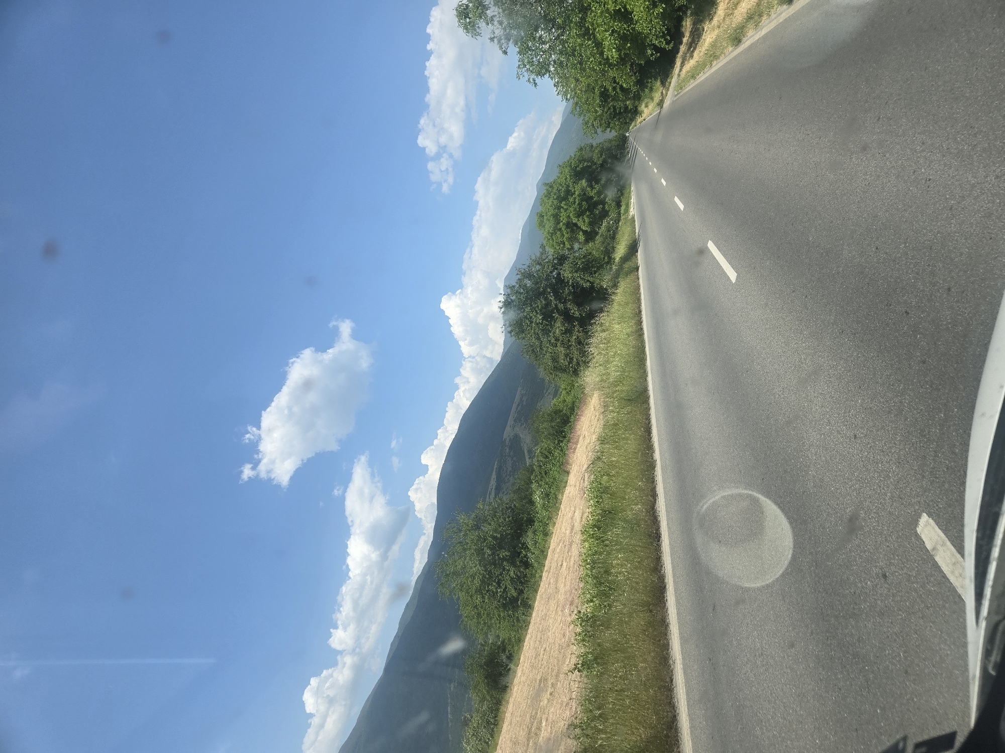 The image shows a view from inside a vehicle, likely a car, driving on a road. The road is asphalt and has white lines marking the lanes. The landscape to the left of the road features a mountain range under a blue sky with fluffy white clouds. Vegetation is present along the roadside, including grass and trees. The image seems to have been taken during the daytime. The specific location (city) cannot be determined from the image alone.