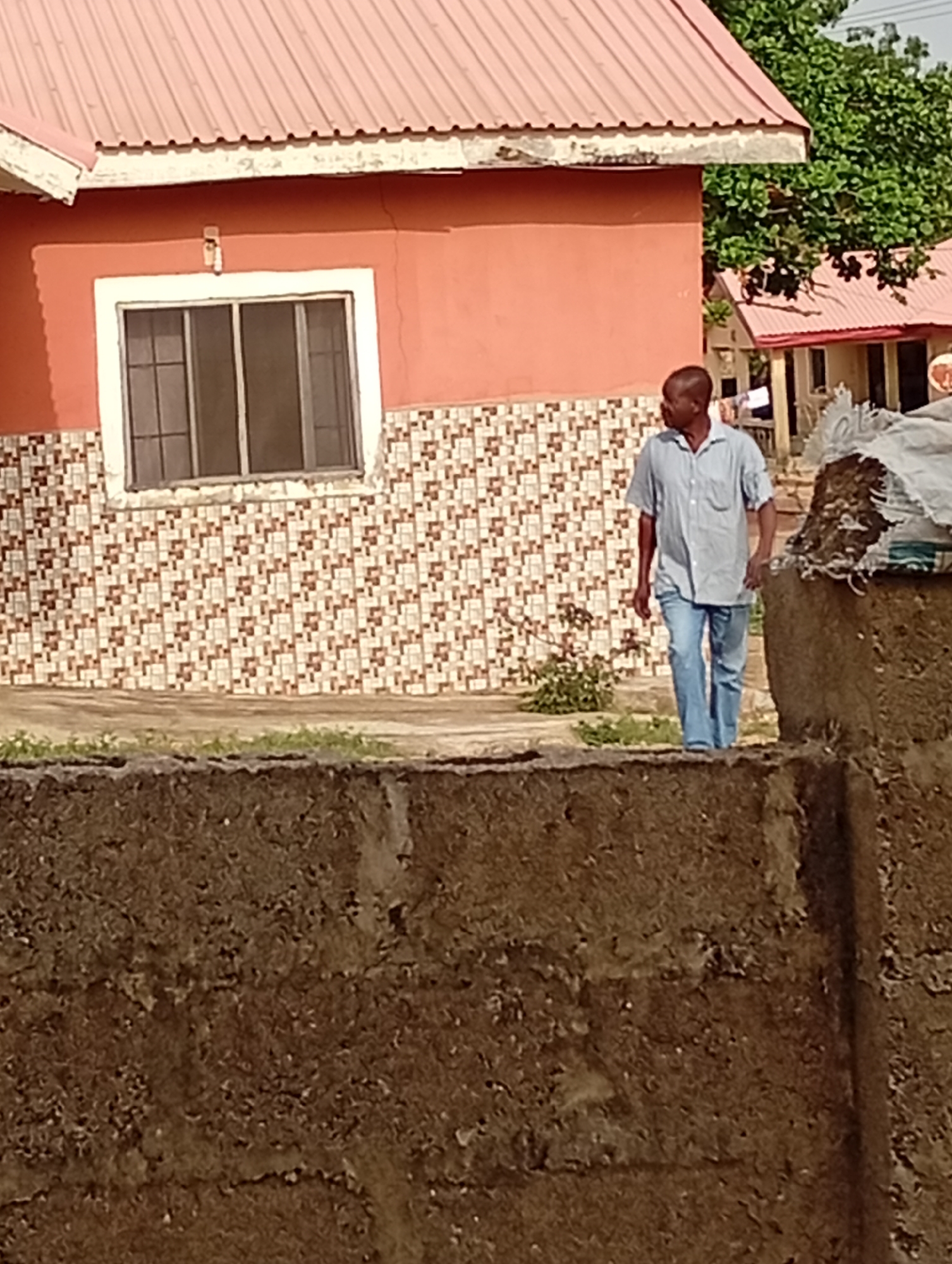 A man wearing a light blue short-sleeved shirt and blue jeans is walking on a concrete wall in Adankolo, Nigeria. Behind him is a salmon-colored building with a reddish-brown corrugated metal roof. The building has a window with a white frame and a tiled base. To the right of the man is another building with a reddish-brown roof, partially obscured by foliage. A white sack sits on the wall to the right of the man. The wall is made of dark brown concrete.