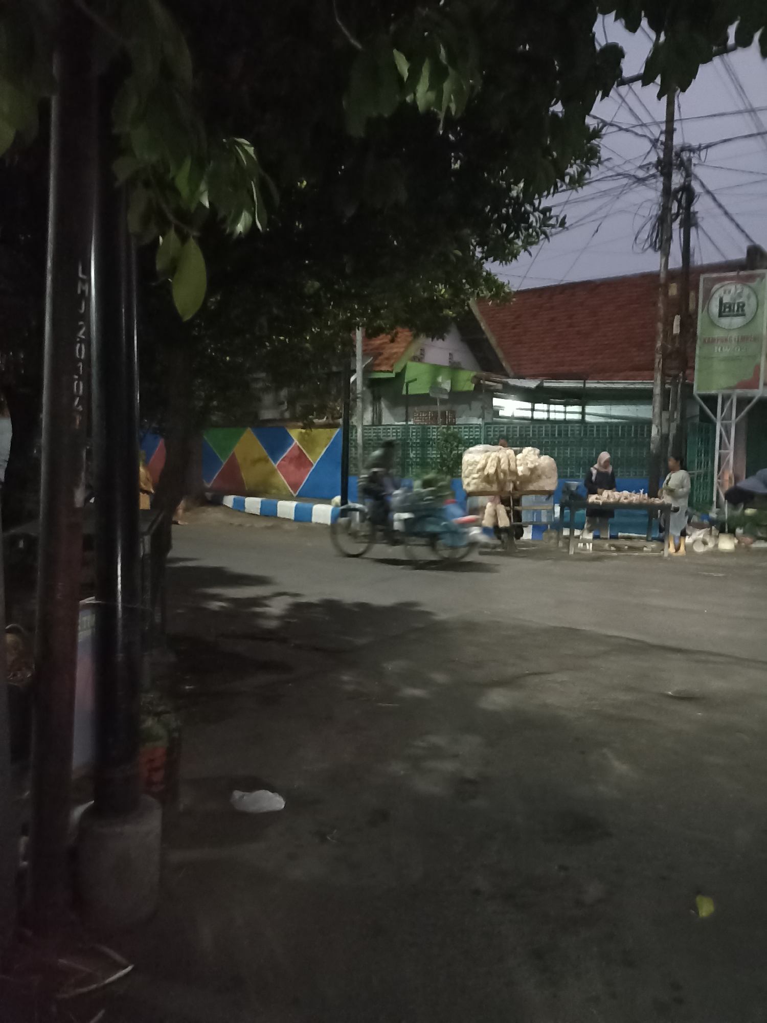 The image depicts a street scene in Ditotrunan, Indonesia, at dusk. In the foreground, a dark, cylindrical pole with vertical white markings is visible on the left. A paved road occupies the majority of the scene, with some scattered debris and shadows.
Further into the scene, a colorful mural adorns a wall on the right side of the road, featuring geometric patterns in blue, yellow, pink, and white. A motorbike with a rider is moving across the road, blurred due to motion.
On the right side of the street, a market stall is set up. Two individuals are present: one, wearing a headscarf, appears to be interacting with items on a table; the other, standing nearby, is looking down. Large, bulky items, possibly produce, are stacked on a cart or table behind them.
A building with a red-tiled roof and green siding is visible in the background. Overhead, a dense network of electrical wires crisscrosses the sky, supported by poles. A sign with the letters "HIR" in a circular emblem is affixed to a pole. The overall lighting suggests it is either early morning or late evening.