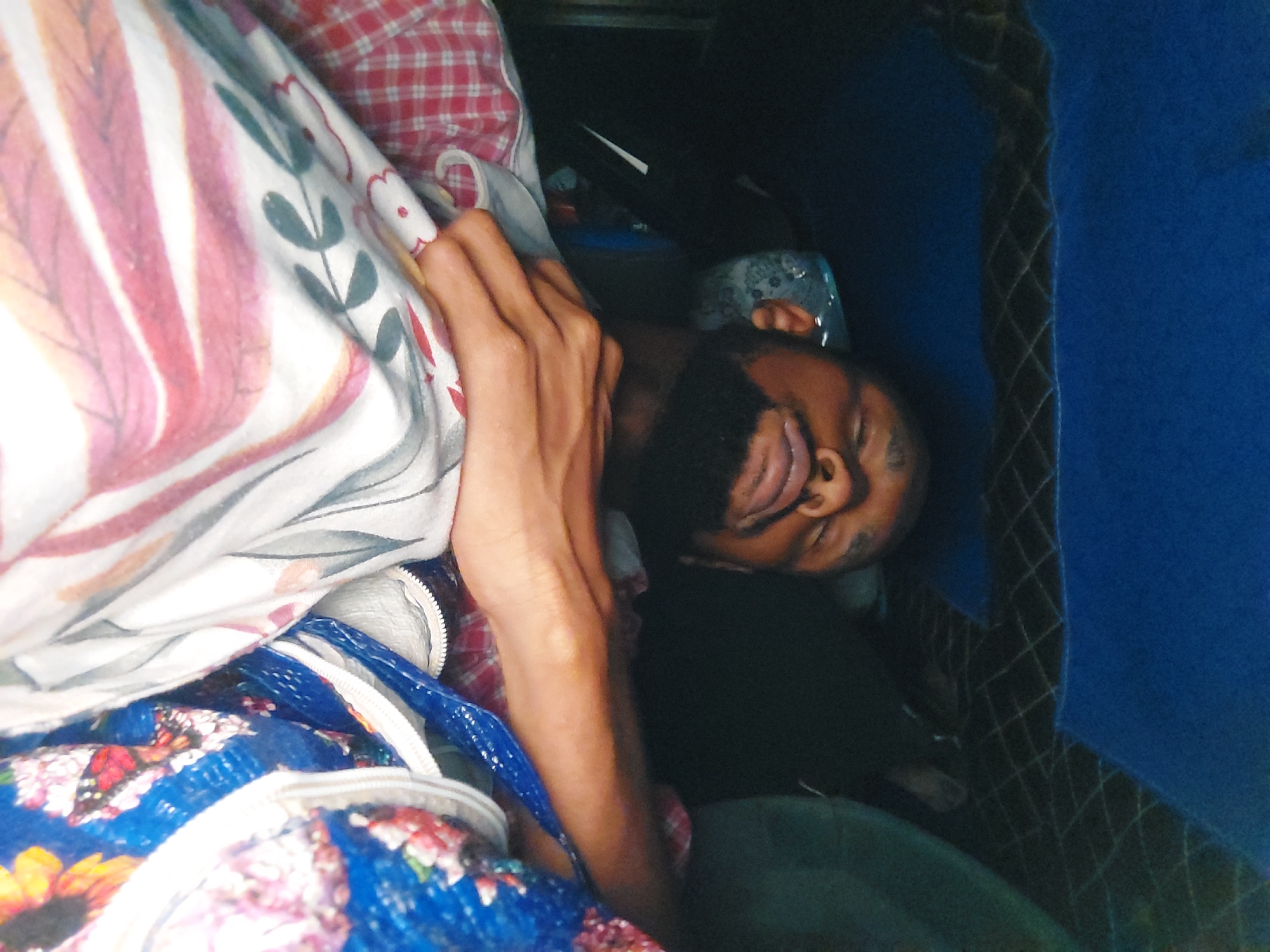 A man with dark skin, a full black beard, and dark hair is lying on his back with his eyes closed and tongue sticking out. He is surrounded by various items including a white and pink patterned blanket, a red and white checkered shirt, and a blue floral patterned bag. He is lying on a blue surface that appears to be fabric. The location is indeterminable and unknown with the provided images.