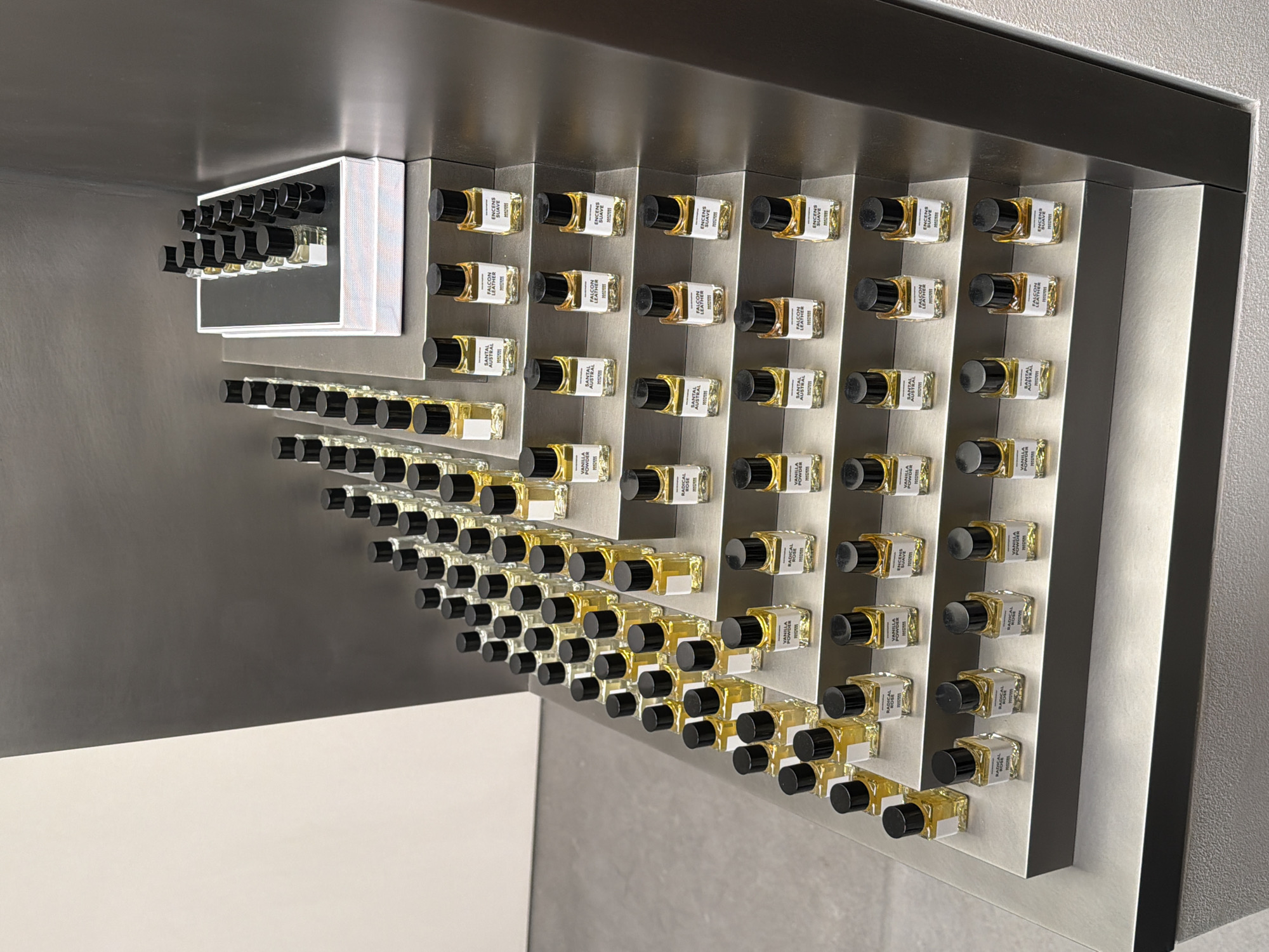 The image depicts a static, elevated view of a fragrance display within a modern retail environment, inferred to be Harvey Nichols in Knightsbridge, London, United Kingdom. Approximately 100-120 small, rectangular glass bottles, each containing a yellow liquid and capped with a matte black screw lid, are meticulously arranged on a multi-tiered display unit constructed from brushed metallic material. Each bottle features a white label displaying fragrance names such as "VANILLA POWDER," "SANTAL AUSTRAL," "FALCON LEATHER," "ENCENS SUAVE," and "RADICAL ROSE," consistently branded "POSSESSIONS." A white rectangular box on an upper tier contains a subset of these bottles. The immediate background consists of a vertical bi-color wall, with a light grey section on the left contrasting with a dark grey section on the right. The floor is a flat light grey. No persons or dynamic actions are present.