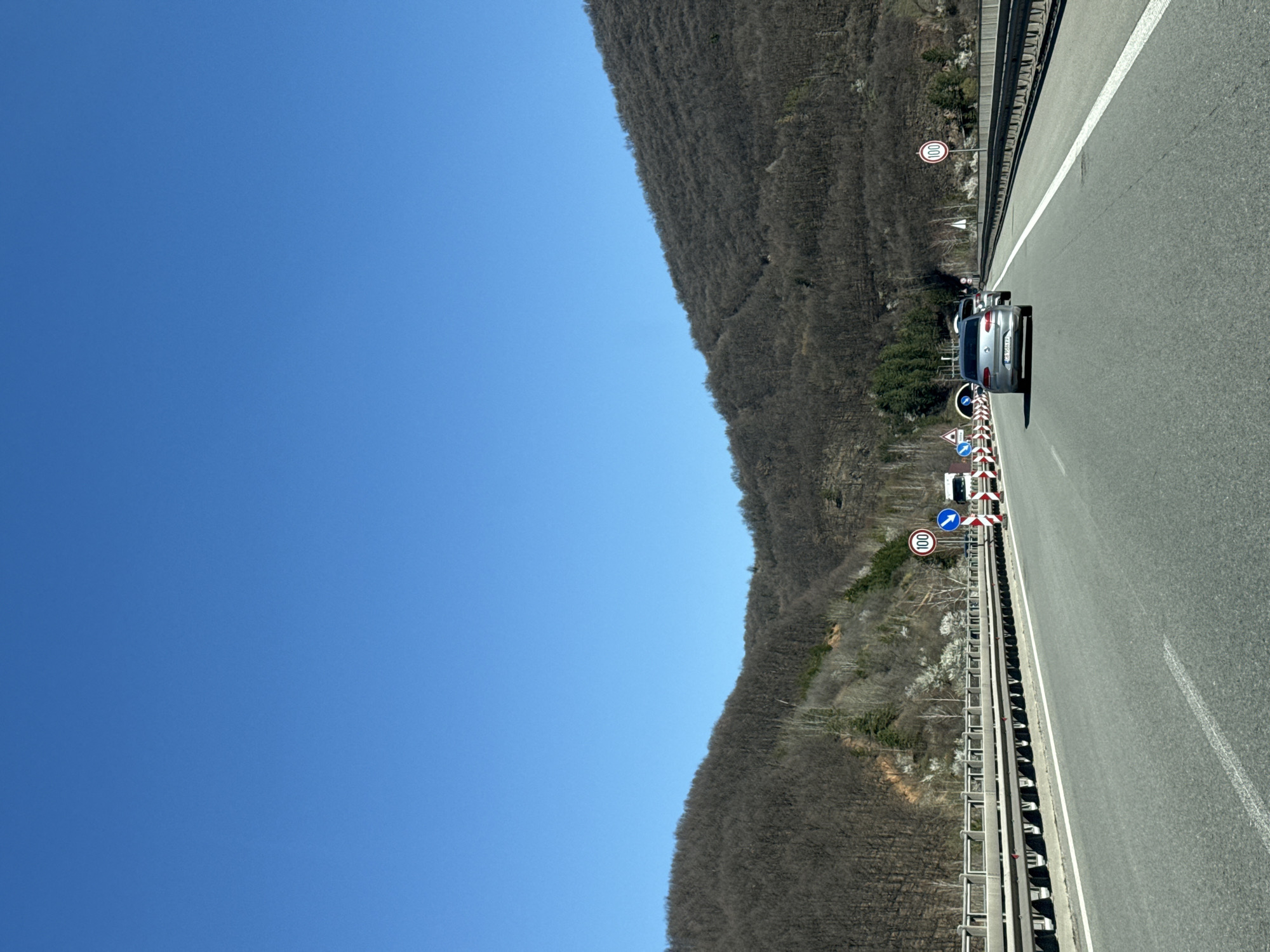 The image shows a highway in Vrachesh, Bulgaria. A silver car is driving on the right lane. There are several road signs and barriers along the side of the road, indicating a possible construction or maintenance area. A forested hillside is visible on the left side of the road, under a clear blue sky.