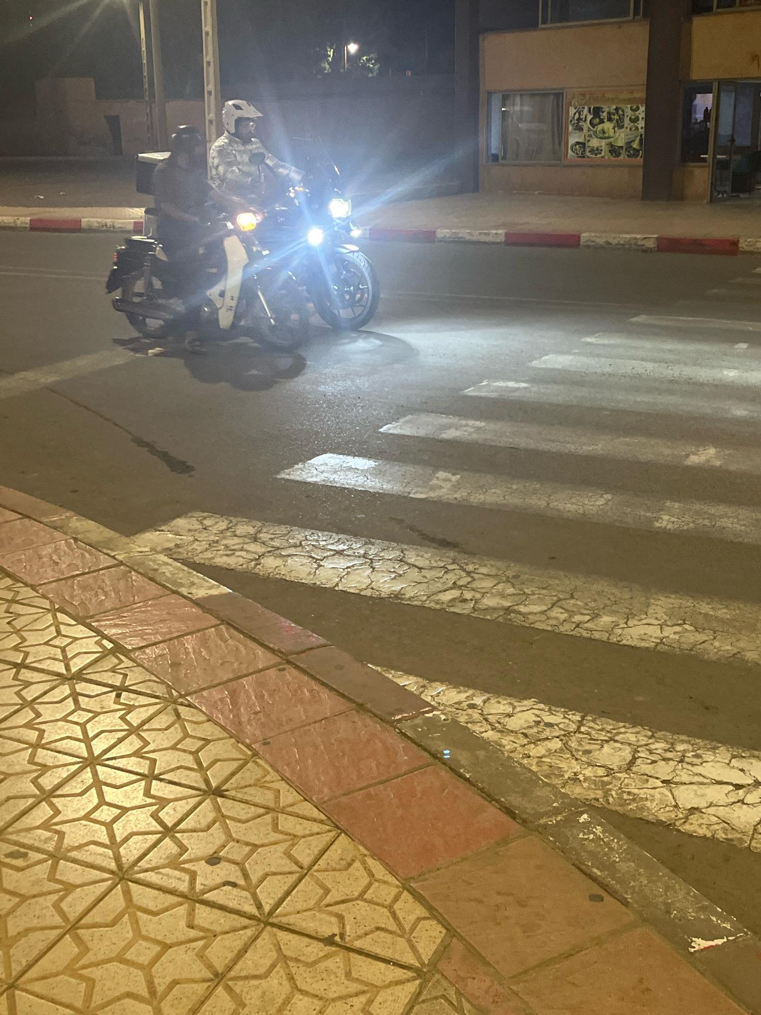The image depicts a nighttime street scene in Azilal, Morocco, focusing on a crosswalk and the adjacent sidewalk. In the foreground, a patterned tiled sidewalk, primarily in yellow with a red border, lines the street. The street itself is asphalt, with prominent white crosswalk markings that appear worn and cracked.
Dominating the midground is a motorcycle with two occupants, illuminated by its bright headlights, which are pointed directly towards the camera, causing lens flare. The driver is wearing a camouflage-patterned jacket and a white helmet, while the passenger behind them is wearing a dark jacket and a dark helmet. The motorcycle has a large, dark box mounted on the rear. The vehicle is positioned on the street, partially obscuring the crosswalk.
In the background, a building with a light-colored facade and darkened windows is visible. A red and white striped curb separates the sidewalk from the street. The overall lighting suggests it is nighttime, with the motorcycle's headlights being the primary light source in the immediate vicinity. The environment appears to be an urban or semi-urban setting.