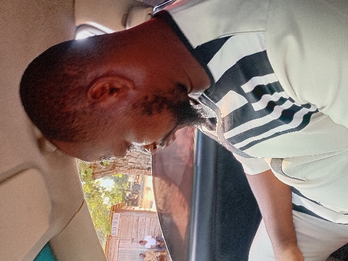 A man with dark skin and a short beard is looking down. He's wearing a white shirt with black and white stripes on the upper portion. The image seems to be taken from inside a vehicle, as parts of the interior are visible. Outside the window, a wooden structure with "BLOCK FOR SALE" written on it is seen, along with a few people gathered nearby. The lighting suggests it could be daytime.