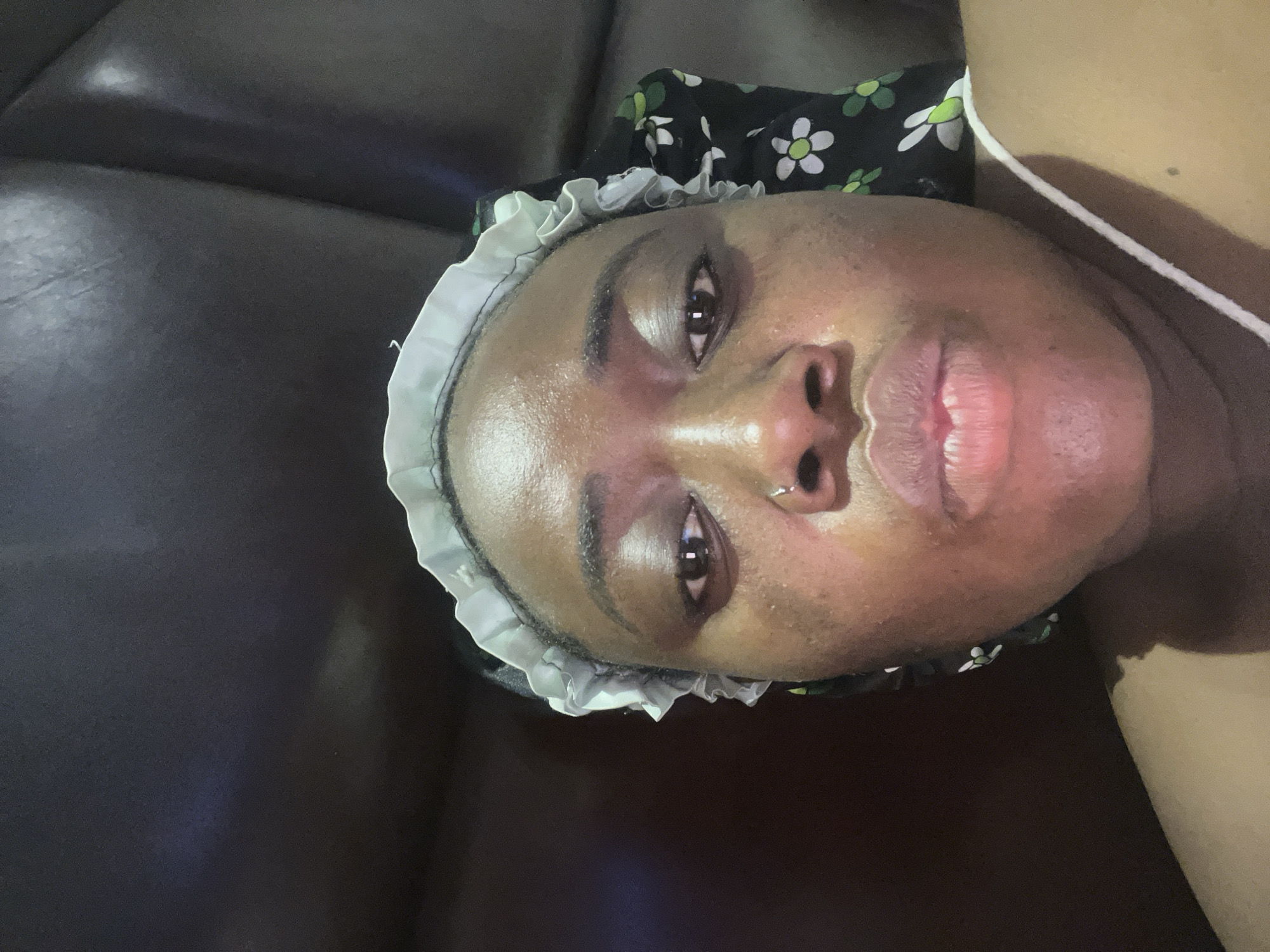An eye-level, close-up shot depicts a dark-skinned person lying on what appears to be a dark, leather-like surface, potentially a couch or similar furniture. The person is wearing a hair covering adorned with a white, frilly edge and a black fabric with a white floral pattern on top. A white strap is visible on the right side of the image near the person's neck and shoulder. The person's face is the primary focus, showcasing smooth skin that seems to be freshly moisturized. There is a piercing in the left nostril. The location is unknown but the user mentions that the image was taken in Abuja, Nigeria.