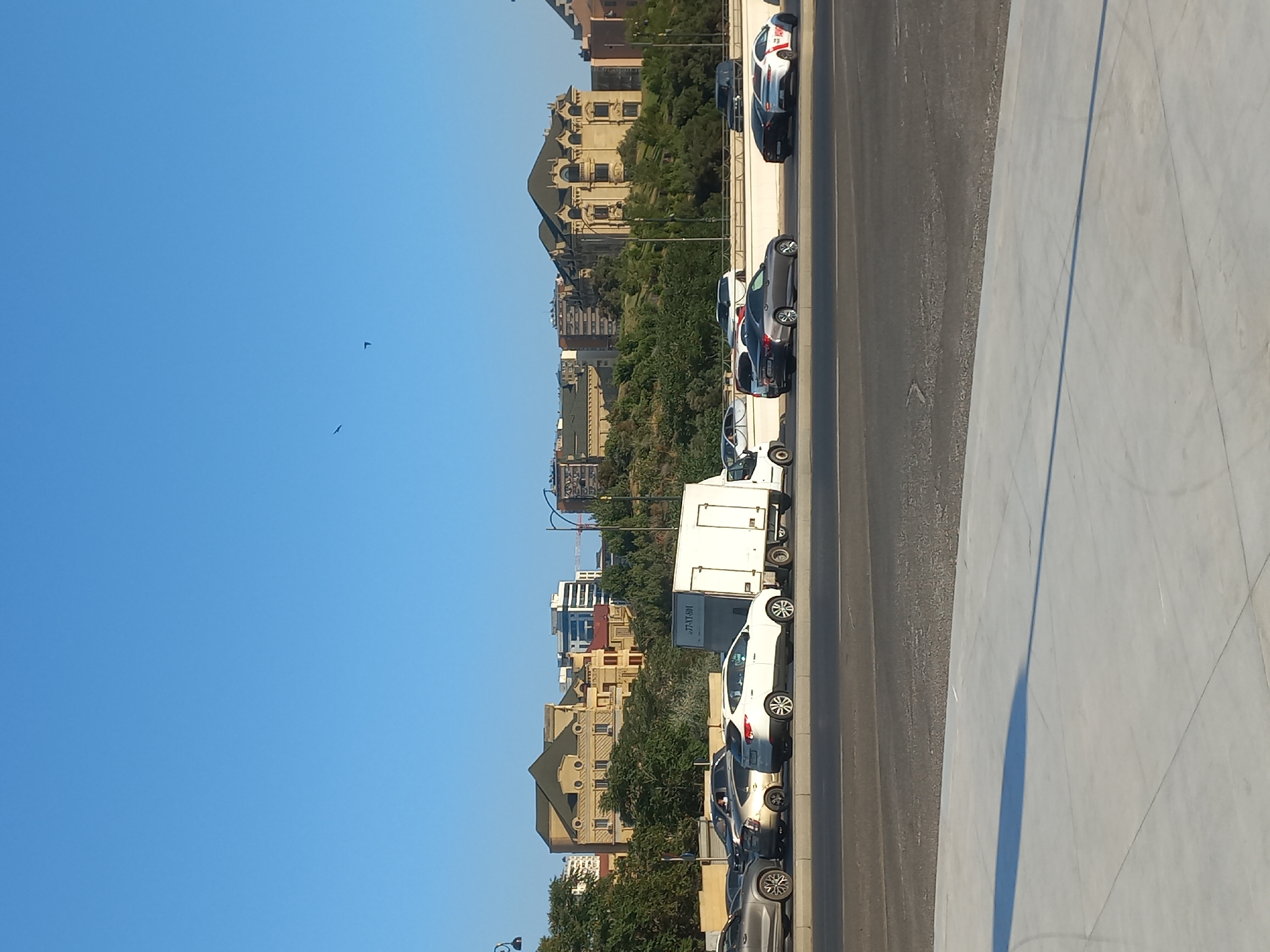 In Baku, Azerbaijan, a street scene unfolds under a clear blue sky. A line of parked cars, including a white truck, a white SUV, and several sedans, are visible on the right side of the road. The buildings in the background are a mix of modern and older architecture, with trees interspersed among them. The foreground features a grey asphalt road and a light-colored concrete walkway, casting a long shadow from an unseen object, likely a lamppost, which is partially visible on the far right. The scene appears to be a typical urban environment on a bright day.