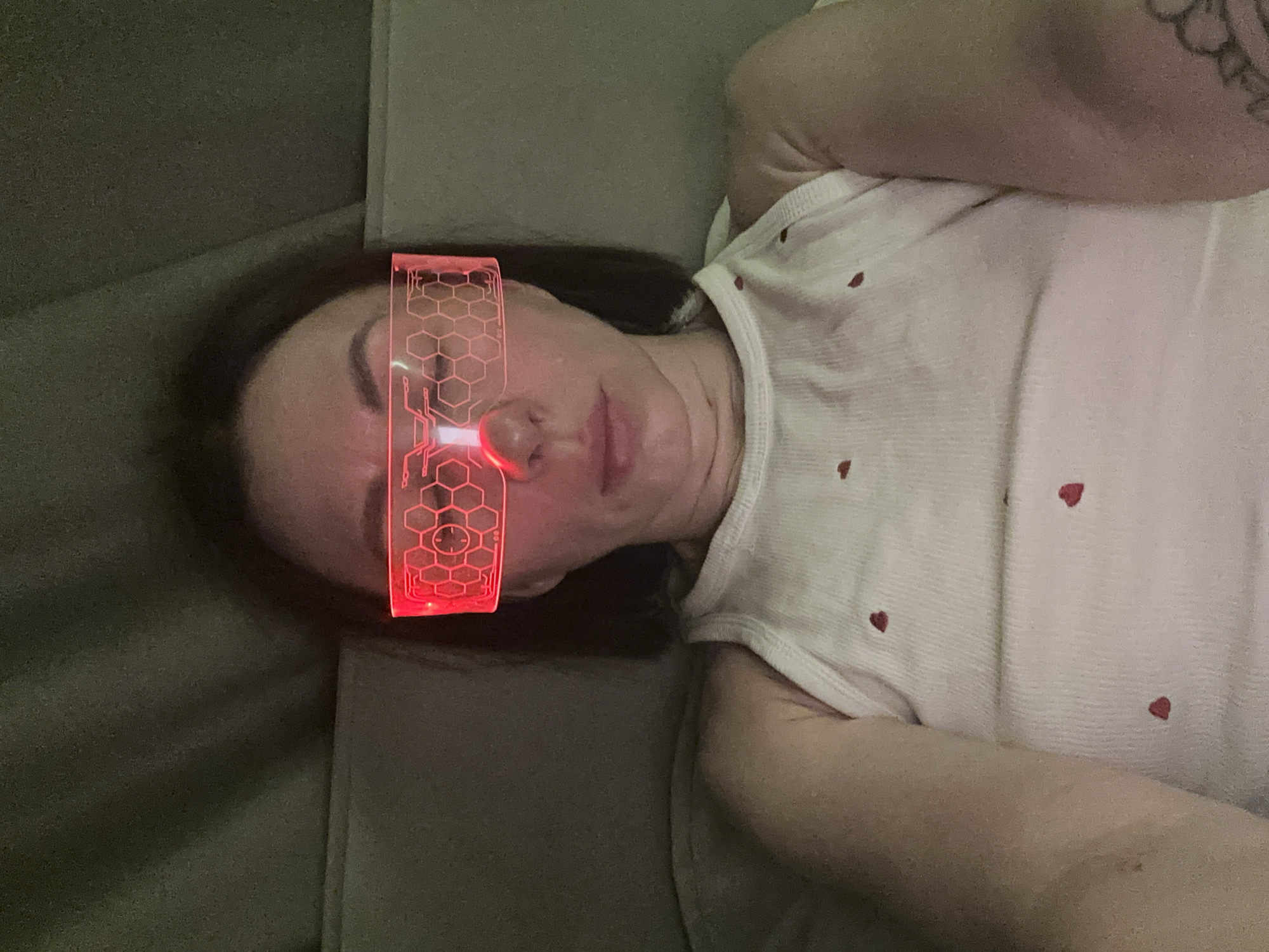 A medium-shot, overhead view shows a person lying on a grey upholstered surface. The person is likely female and has dark hair. They are wearing a white tank top with small red heart patterns. Their left arm is bent upwards toward their face. The person is wearing a pair of red, illuminated, novelty glasses with a honeycomb and spiderweb pattern. The glasses are positioned over their eyes, partially obscuring their face. A tattoo is visible on the upper right arm. The background is a grey, textured surface, likely part of the furniture the person is lying on. The location is not evident from this image.