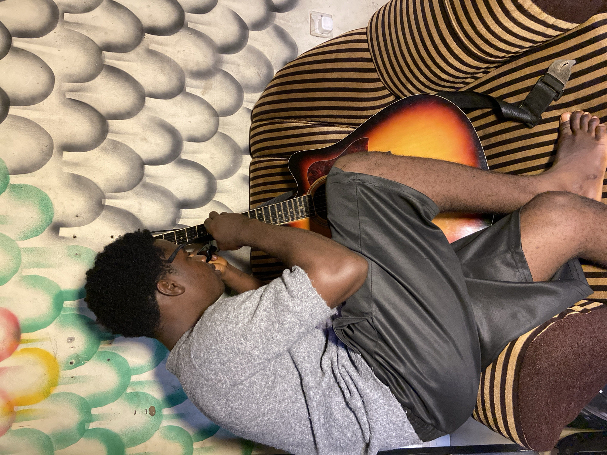 An overhead, slightly angled shot captures a young adult male lying on his side and playing an acoustic guitar indoors, likely in Mararaba, Nigeria. The individual, with dark skin and short, dark curly hair, wears glasses, a light grey short-sleeved shirt, and dark shorts. He appears relaxed, resting his upper body against a wall and his lower body on a striped sofa. His right hand is positioned on the guitar's fretboard, while his left hand is near the soundhole, engaged in playing. His bare feet are visible, one resting on the striped upholstery of the sofa.
The acoustic guitar features a sunburst finish, transitioning from dark orange/red to a brighter yellow. A black strap is draped over the sofa cushion near the guitar's body. The background wall is decorated with a painted pattern of overlapping, scale-like or bubble shapes, predominantly in shades of grey and white. Towards the bottom left, the pattern incorporates vibrant green and hints of other colors. Above the sofa, a white electrical outlet is affixed to the patterned wall. The sofa itself is upholstered in a distinctive brown and light brown/gold striped fabric. There is no visible text in the image.
