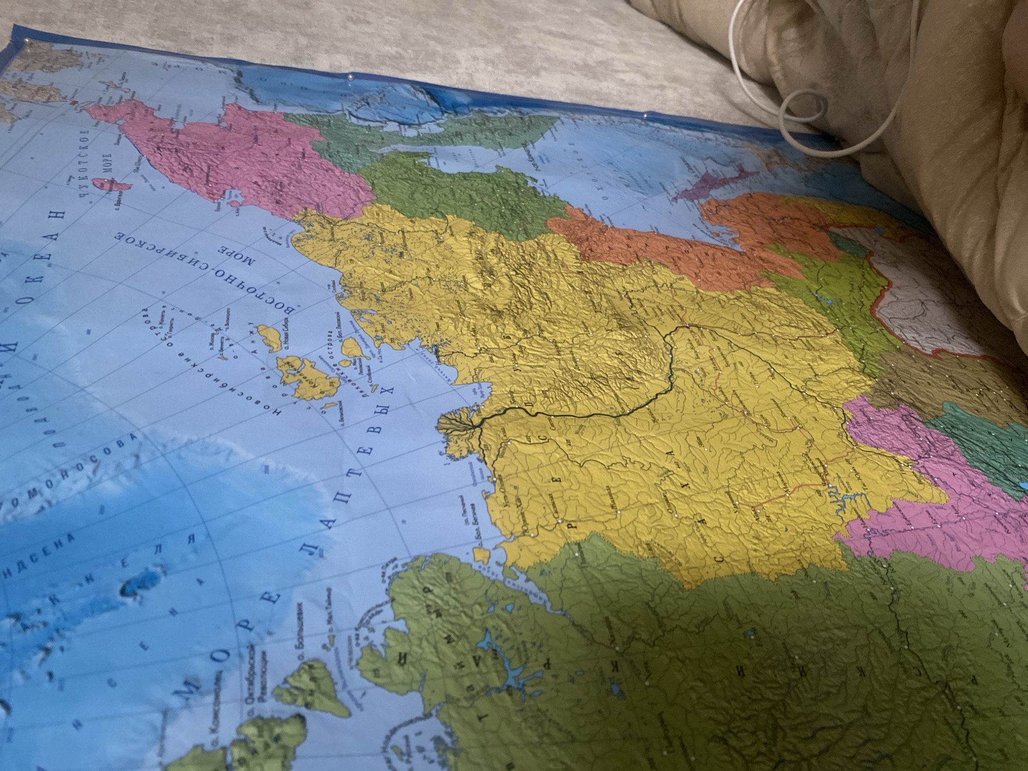 The image shows a close-up, angled view of a large wall map of Russia. The map is colorful, with different regions highlighted in various colors like green, yellow, pink, and orange. Text and place names are visible in Russian on the map. The map is attached to a wall with what appear to be small pins or magnets on the right side. The environment surrounding the map is out of focus but suggests an indoor setting, possibly a room with soft furnishings, indicated by the presence of a beige fabric texture in the foreground and background. There are no people visible in the image. The specific location of Nikologory, Russia, is not depicted or identifiable within the visible portion of the map.