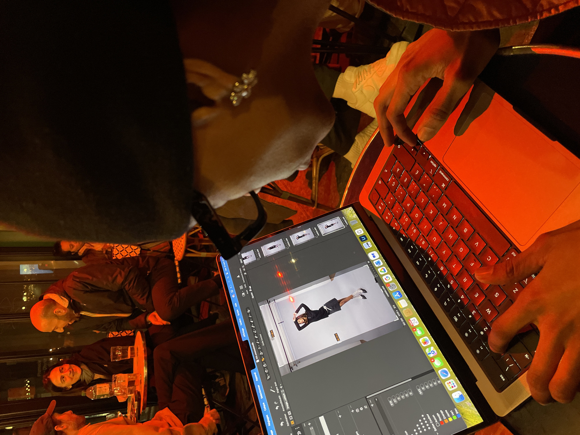 A person is depicted from a high angle, focused on a silver MacBook laptop with a red backlit keyboard and trackpad. Their hands are positioned over the keyboard and trackpad. The individual wears a dark beanie, black-rimmed glasses, and an ear piercing with multiple small beads on their left earlobe. The laptop screen displays an image editing application (likely Adobe Photoshop, with a French interface), featuring a full-body studio portrait of a person posing in a black long-sleeve top, black mini skirt, white socks, and black shoes. Multiple thumbnail images are visible on the right side of the screen.
In the background, three individuals are seated at a small round table in a dimly lit cafe or restaurant environment. From left to right, a male in a reddish-orange shirt and a baseball cap is partially visible, facing a smiling female in a dark jacket and scarf. She is looking towards a bald, bearded male in a dark puffer jacket who is speaking. A clear bottle and two glasses are on their table. Bistro-style chairs are visible. Reflections on a glass partition behind them reveal text, including "Pharmacy" and "Oberkampf," identifying the location as a cafe or restaurant on Rue Oberkampf in Paris, France, at night. The foreground, including the laptop and the user's hands, is illuminated by a prominent red light source.