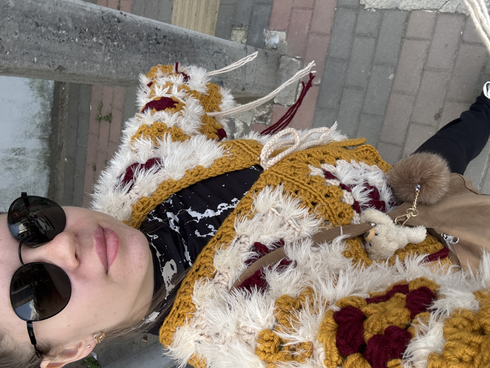 A person, presumably a young woman, is the primary subject of the image, taken in an elevated, tilted, close-up selfie perspective. She is wearing black sunglasses, and her face is partially visible, showing fair skin and pink lips. She is adorned with a large, colorful, crochet-style coat, with a pattern of brown, white, and maroon hues, suggesting a hand-crafted garment. Part of a black and white patterned garment is visible beneath the crochet coat. Beside her is a suede-like jacket. There is a furry keychain with a small stuffed animal and a chain that is clipped to the jacket.
The background reveals a street scene, identifiable by the presence of a grey concrete barrier or wall on the left side and a pedestrian walkway paved with multicolored bricks or stones in the upper right corner of the image. The location appears to be a street or sidewalk, with a hint of urban environment visible.
While specific elements might suggest Batumi (e.g., architectural style or unique local features), there is no direct visual evidence to confirm this location. Without additional information or landmarks visible in the image, the precise location cannot be definitively determined.