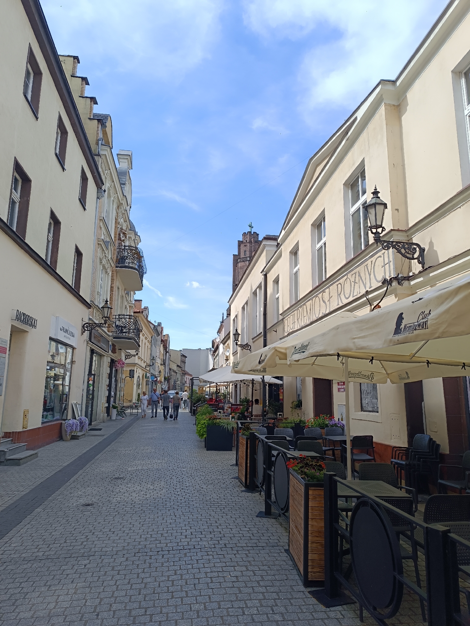 The image depicts a street scene in Gliwice, Poland, likely on a pleasant day with a blue sky and scattered clouds. The street is paved with cobblestones. On the left side of the street are buildings with storefronts, including "RACHORSKA" and "FRANCISZKA". Several people are walking down the street. On the right side of the street, there are outdoor seating areas for cafes or restaurants, with tables and chairs set up under large umbrellas. The architecture of the buildings appears to be historic. The overall atmosphere is that of a European city street.