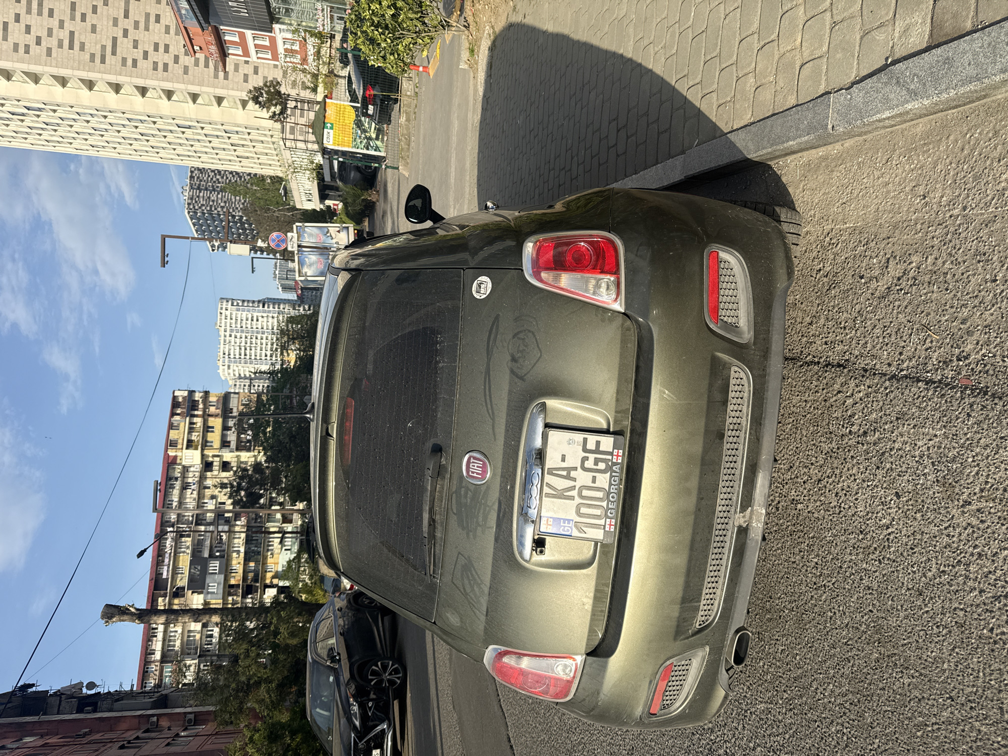 The image depicts the rear of a green Fiat 500 parked on a street in Batumi, Georgia. The license plate number is KA-100-GF. The car is parked alongside a brick sidewalk. Buildings are visible in the background, including high-rise residential apartments and other commercial structures. The sky is clear and blue. A black car is parked on the road behind and to the left of the Fiat.