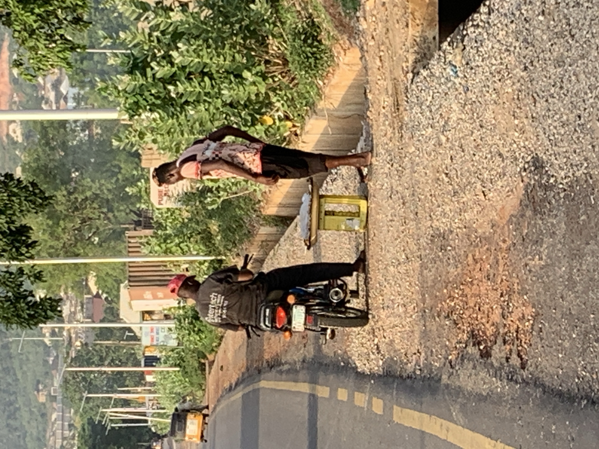 In Zango-ndaji, Nigeria, a man wearing a black shirt and a red cap sits on a parked motorcycle on the side of a paved road. A second man in a floral shirt stands on the dirt next to a yellow crate. Trees and buildings are visible in the background.