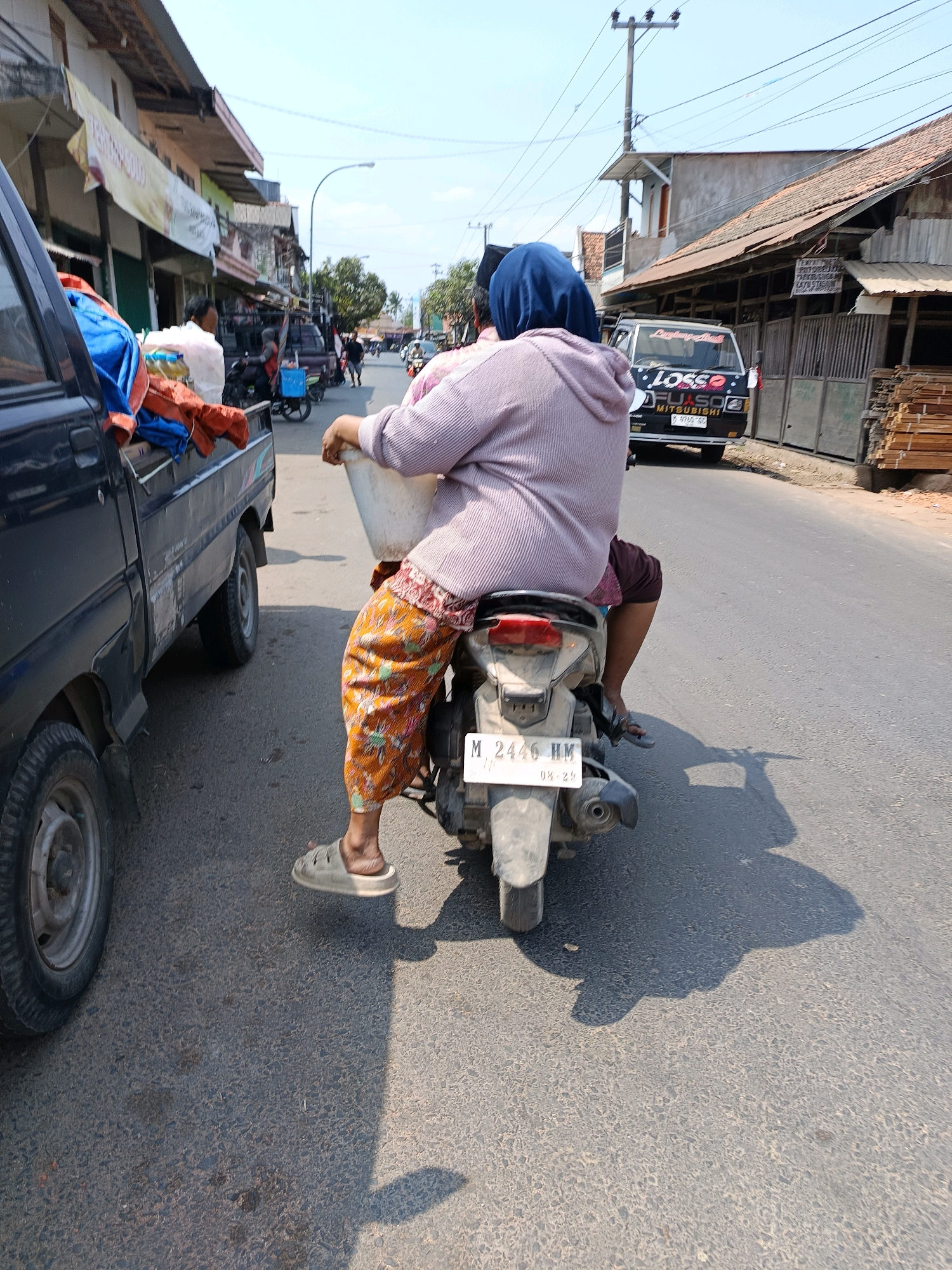 The image shows a street scene in Blega, Indonesia. A motorcycle, with two passengers, is traveling on the right side of the road, next to a black pickup truck. The passenger in the front, a woman wearing a blue hijab and a light purple cardigan, is holding a white bucket. The passenger behind her is wearing a dark shirt. Both are wearing casual clothing. The pickup truck on the left is loaded with various items, including orange and blue fabrics. The road appears to be asphalt, with some debris visible. Buildings line the street on both sides, with some appearing to be shops or small businesses. A utility pole with power lines is visible in the background. The sky is clear and blue, indicating a sunny day. A van with the word "Losso" and "MITSUBISHI" written on it is parked further down the road on the right. The license plate of the motorcycle reads "M 2446 HM".