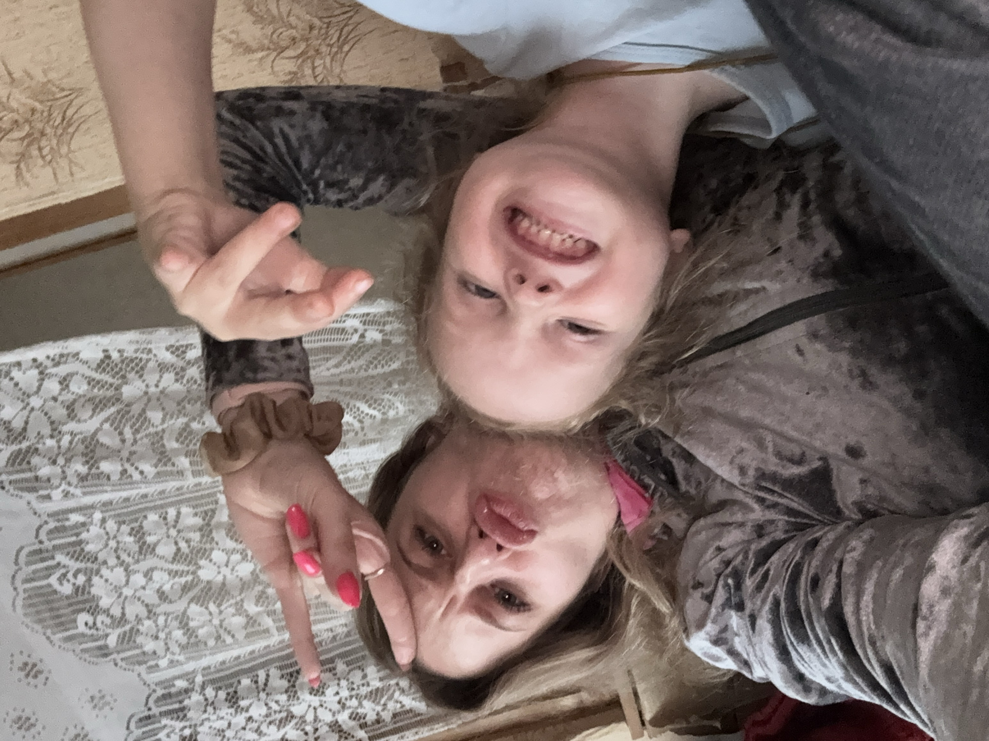 In an indoor setting in Severnyy, Russia, a woman and a young child are captured in a close-up selfie. The woman, with blonde hair and vibrant pink fingernails, is making a duck face and has her index and middle fingers raised in a peace sign, her ring finger adorned with a simple band. She wears a dark, plush velvet top with a leopard-like pattern. To her right, the child, with blonde hair and a playful expression, bares their teeth in a wide smile. The child is dressed in a light blue t-shirt. The background reveals a lace curtain to the left and a textured fabric, possibly upholstery, to the right. The overall scene conveys a moment of shared joy and lightheartedness.