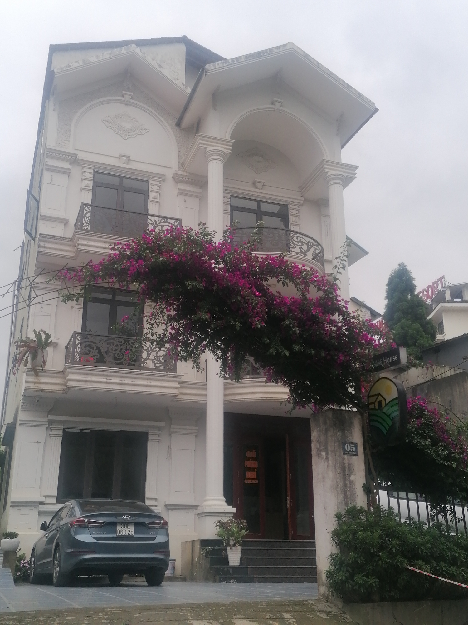 The image captures a multi-story white building with ornate classical architectural features, likely a hotel or guesthouse, under an overcast sky. The building boasts decorative balconies with intricate dark wrought iron railings and prominent white columns on its upper floors. A large, vibrant pink bougainvillea vine cascades dramatically across the front facade, adding a striking burst of color against the white building and muted sky.
Parked in front of the ground floor entrance is a dark blue Hyundai Elantra sedan, bearing the Vietnamese license plate "30E 060.25". The car's model "ELANTRA" and trim "GLS" are visible on its rear. The building's main entrance features dark wooden double doors and a short flight of steps. To the right of the entrance, a concrete pillar displays the number "05" and the text "PHO." Another sign, a circular one depicting a stylized house on green hills, hangs nearby, partially integrated into the bougainvillea. In the background, past the building and some trees, another structure with a sign partially visible as "ESORT" (likely "RESORT") can be seen.
The sky is a uniform, pale gray, indicating a heavily overcast or misty day, characteristic of Sa Pa. The ground appears damp. There are no people visible, suggesting a quiet moment at the location. The overall scene depicts an elegant building nestled in a lush, cloudy environment.