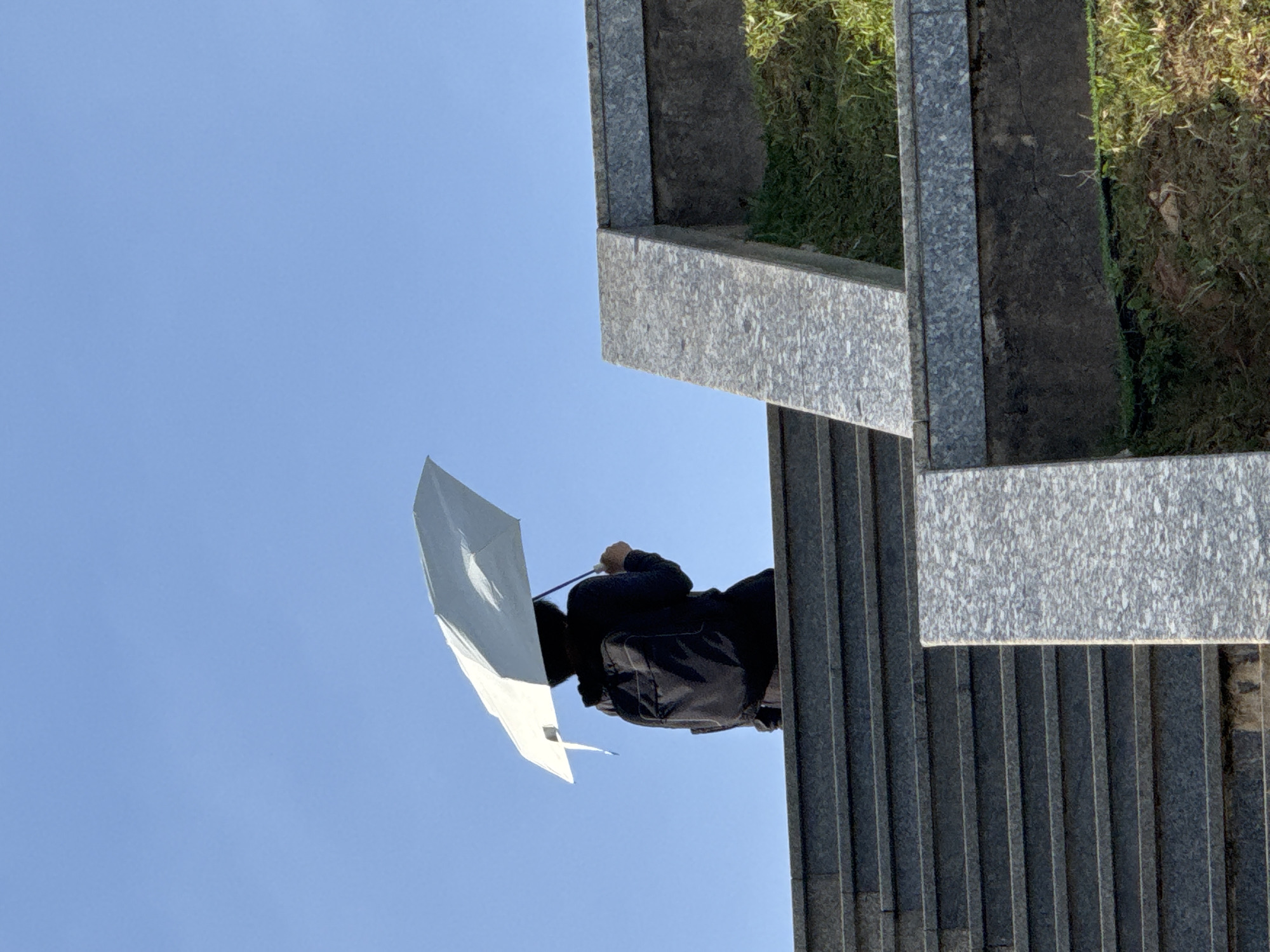 A person wearing a black jacket and a backpack is sitting on a stone staircase and holding a white umbrella over their head. The person is viewed from behind, and their face is not visible. The staircase is made of dark gray stone with lighter gray granite accents. Green grass is growing on the side of the staircase. The sky is clear and blue. The scene is set in Da Lat, Vietnam.