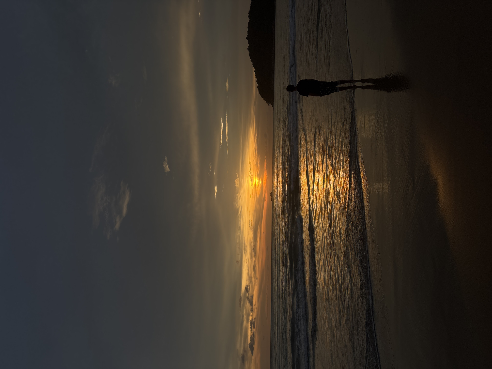 The image shows a beach at sunset in Choeng Thale, Thailand. A silhouetted figure stands at the edge of the water. The sky displays hues of orange and yellow where the sun dips toward the horizon. Dark clouds fill the upper portion of the sky. A land mass is faintly visible on the right side of the image. Waves reflect the sunlight onto the shore.