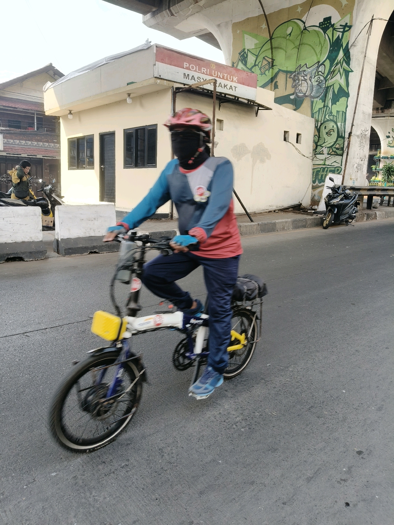 In South Tangerang, Indonesia, a person wearing a helmet, face mask, and a blue and red long-sleeved shirt is riding a folding bicycle. The bicycle has a yellow holder on the front and bags attached to the rear rack. The rider is on a paved road, with a building featuring a sign that reads "POLRI UNTUK MASYARAKAT" (Police for the Community) and graffiti art visible in the background. A motorcycle is parked on the side of the road near the building. Another person on a motorcycle is visible further in the background. The scene takes place under a large elevated structure, likely an overpass or bridge, with the sky visible above.
