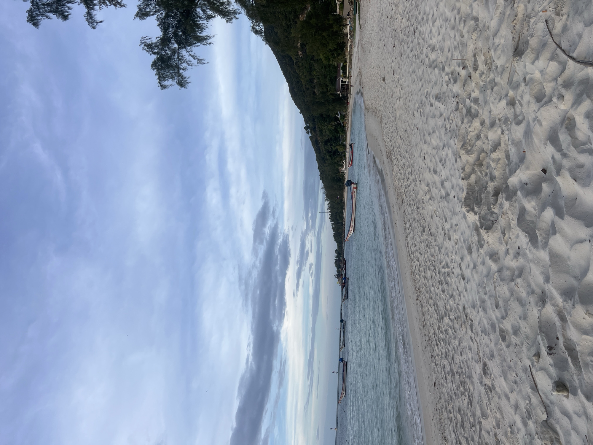 The image depicts a tranquil beach scene under a cloudy sky. The foreground is dominated by soft, white sand marked with footprints and scattered debris. The gentle waves of the turquoise ocean lap at the shore. Several longtail boats are anchored in the shallow water, some with outboard motors. In the background, a lush green hillside covered in trees slopes down to the beach. Small buildings and structures are visible nestled amongst the trees. A lighthouse-like structure stands on a distant point of land to the left. The overall atmosphere is peaceful and serene.