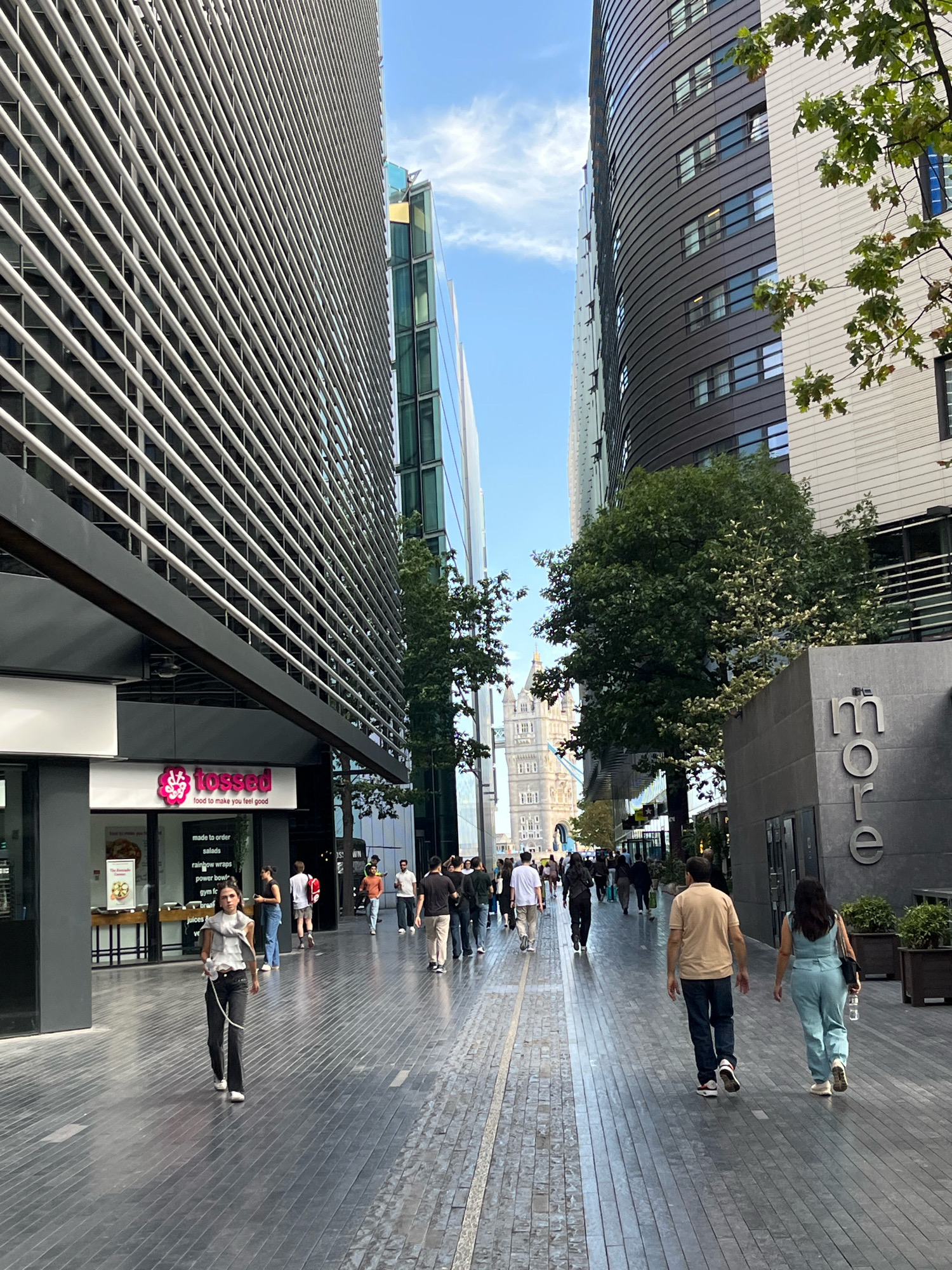 This image captures a bustling pedestrian walkway in London, United Kingdom, with modern architecture dominating the scene. The walkway is paved with dark grey rectangular tiles, reflecting the light from the sky. On the left, a large, sleek building features a facade of horizontal metal louvers, with a "tossed" restaurant visible at its base, displaying its slogan "food to make you feel good." To the right, a curved, dark-colored building with rows of windows rises, adjacent to a lighter-colored, horizontally paneled building. Further down the walkway, a sign with the word "more" in large letters is mounted on a dark grey structure. In the distance, Tower Bridge is visible, framed by trees and the surrounding buildings, under a bright, partly cloudy sky. Several people are walking along the path in both directions, dressed in casual attire. A young woman in the foreground walks towards the camera, holding a leash attached to a dog that is out of frame. Other individuals are seen walking in groups and individually, contributing to the lively atmosphere of the urban environment. Green trees and planters add touches of nature to the otherwise concrete and glass landscape.