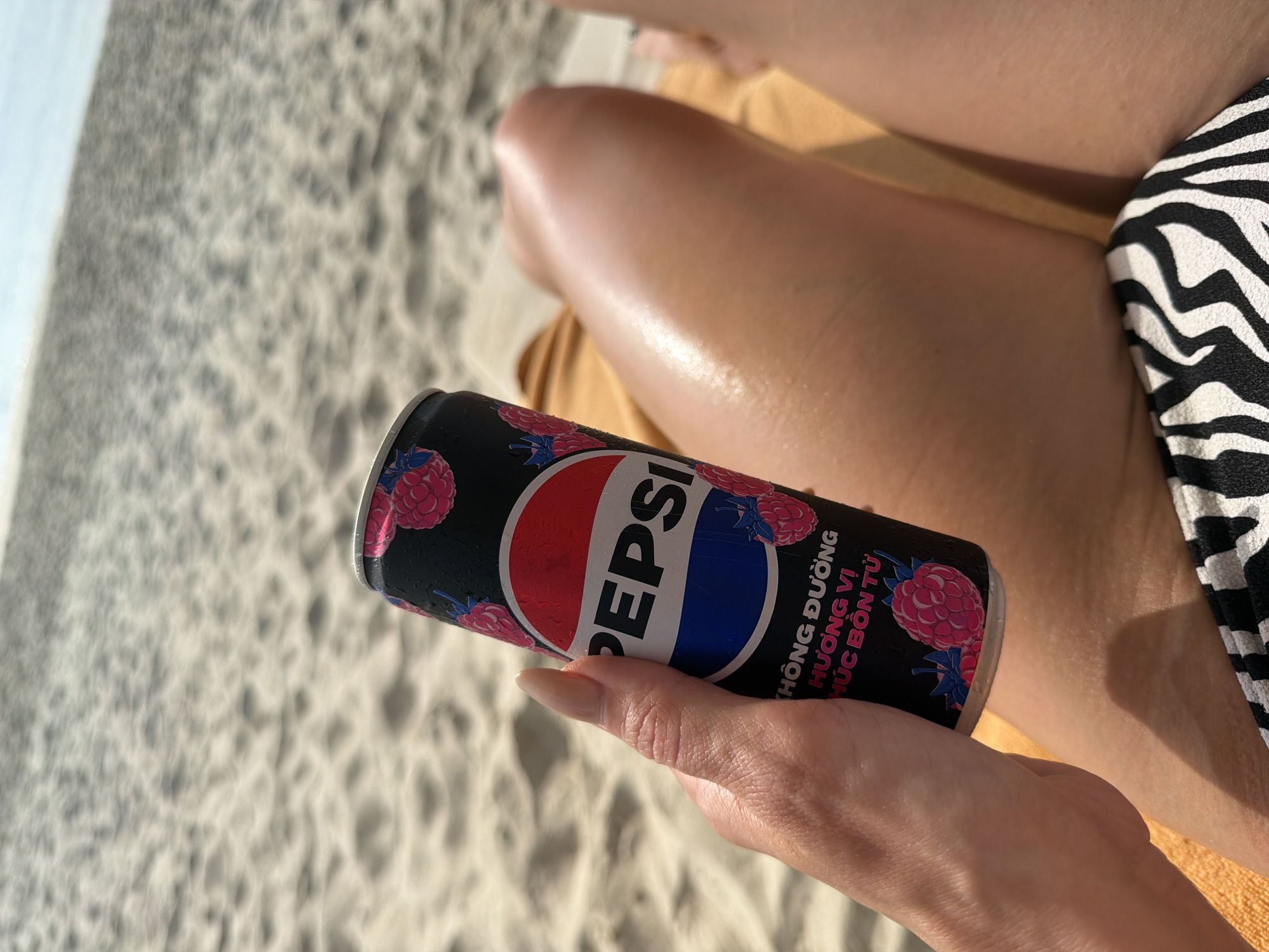 A person's hand holds a can of Pepsi with raspberry imagery on its black exterior. The can has the Pepsi logo in its signature red, white, and blue colors. Text in Vietnamese is visible below the logo. The person is sitting on a beach towel on a sandy beach, with the ocean and sky blurred in the background. The scene suggests a relaxing day at the beach in Ninh Hoa Ward, Vietnam, possibly during a vacation.