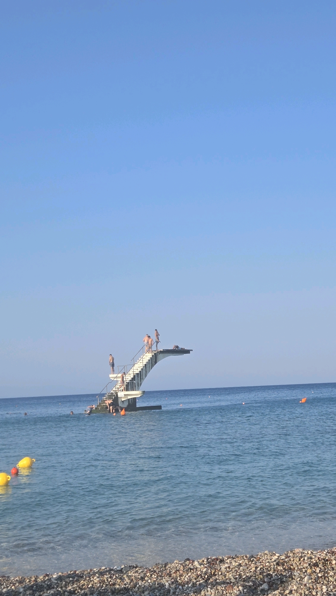 In the Municipal Unit of Rhodes, Greece, on a clear, sunny day, a diving platform stands in the blue sea. The platform has a white, multi-tiered structure with stairs leading up to a diving board. Several people are visible on and around the platform. Some are standing on the diving board, preparing to jump, while others are on the stairs or at the base of the platform. The water is calm, with gentle waves. Yellow and red buoys are floating in the sea. The beach in the foreground is covered with pebbles. The sky is a clear, bright blue.